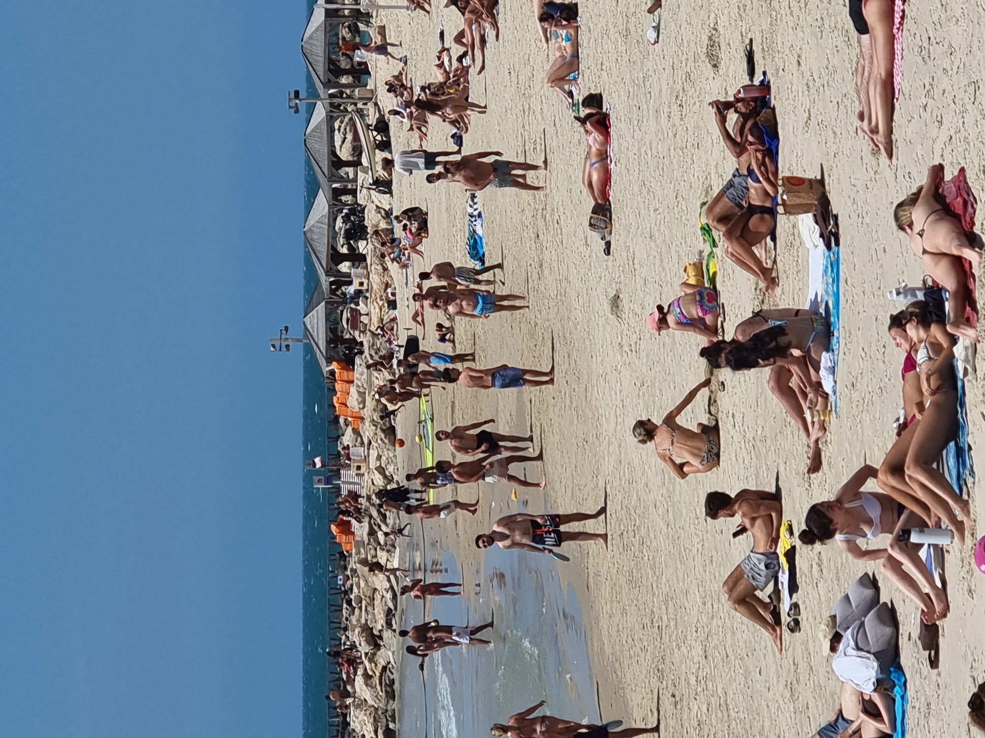 The image captures a crowded beach scene in Tel Aviv, Israel. Numerous people are present, engaging in various activities such as sunbathing, swimming, walking along the shoreline, and playing beach games. The beach is sandy, and the sea stretches out to the horizon under a clear, blue sky. To the left, a stone breakwater is visible, lined with orange buoys and additional beachgoers. Beach umbrellas and shelters are sparsely scattered among the people. The scene suggests a typical sunny day at a popular coastal destination.