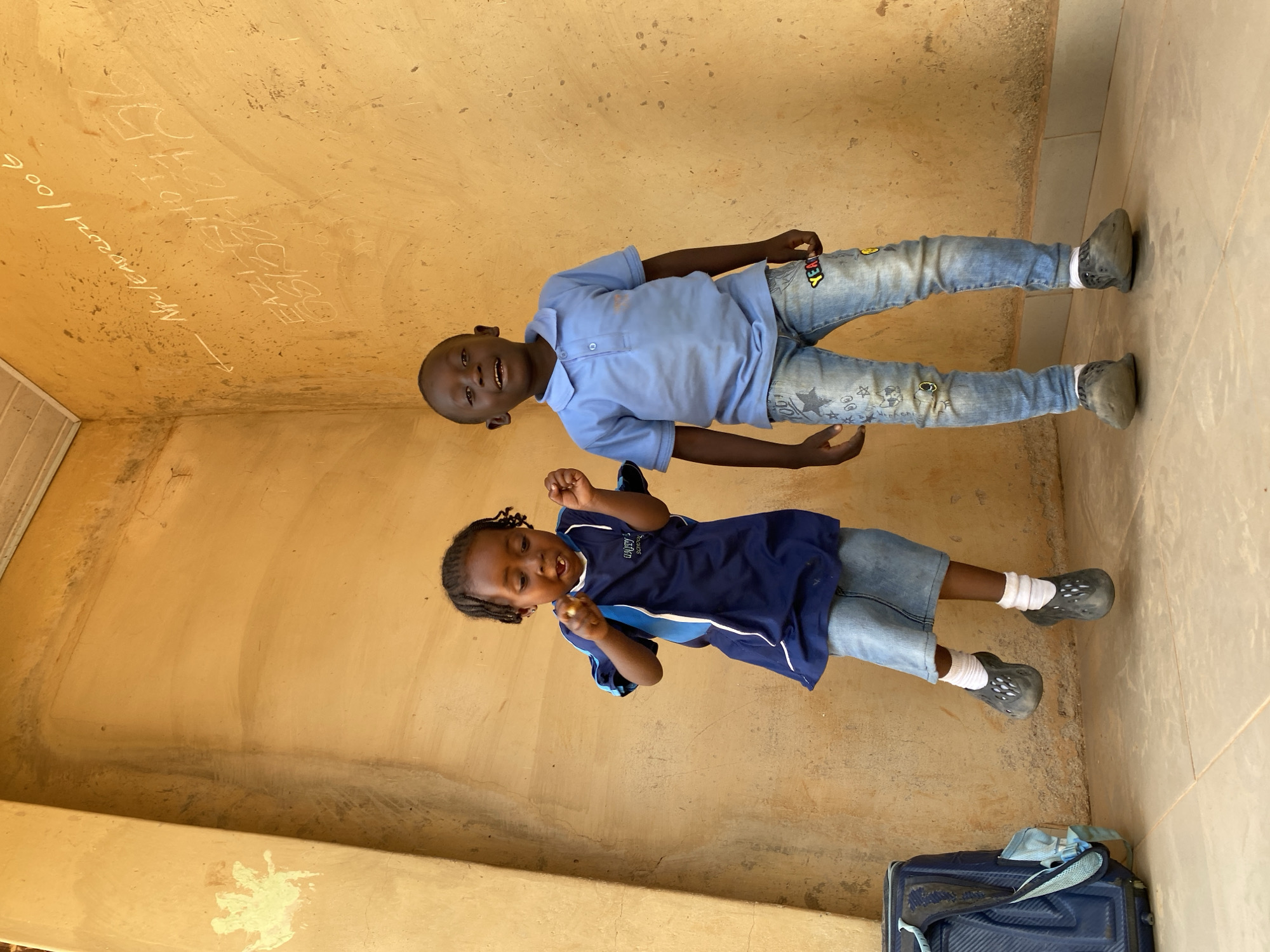 Two young boys are positioned indoors, seemingly lying on their sides against a light tan wall and beige tile floor, possibly in a room within a building in Kuru Dungus, Nigeria. The older boy wears a light blue polo shirt and patterned denim jeans with grey slip-on shoes. The younger boy wears a dark blue and light blue shirt with denim shorts and grey slip-on shoes with white socks. An arrow and faded writing are visible on the wall. A blue suitcase sits on the floor.
