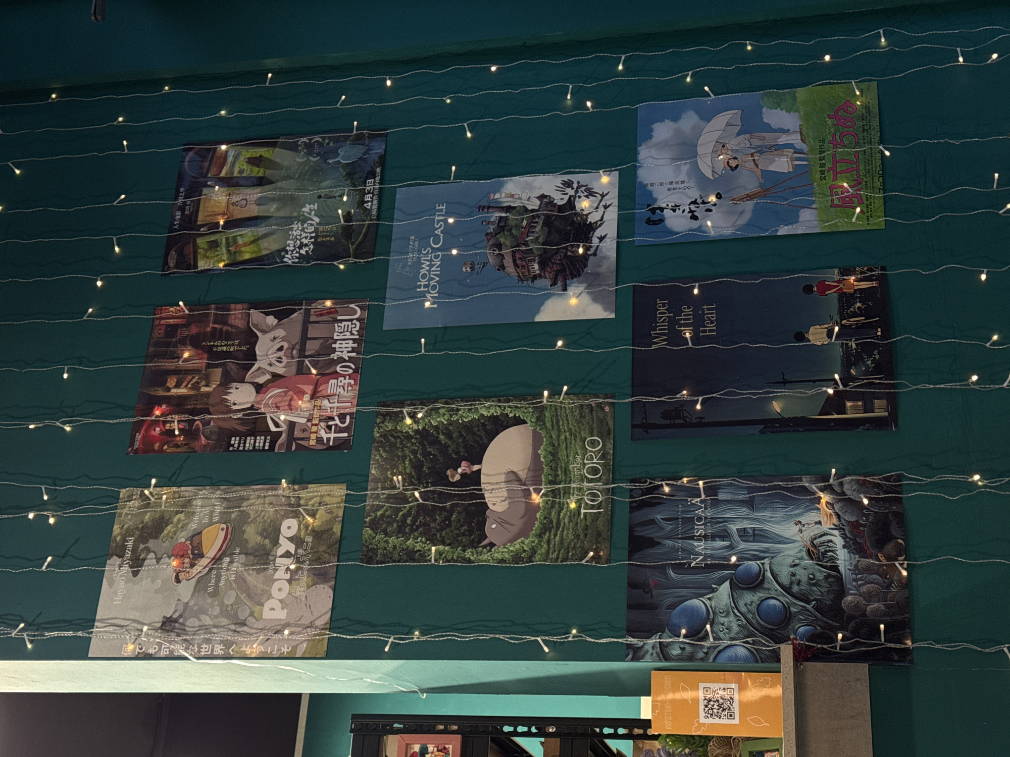The image shows a green wall adorned with fairy lights and nine Studio Ghibli movie posters arranged in a 3x3 grid. The posters include "Howl's Moving Castle," "My Neighbor Totoro," "Ponyo," "Spirited Away," "Whisper of the Heart," "Nausicaä of the Valley of the Wind," "The Wind Rises," and "How Do You Live". The bottom portion of the image reveals a shelf with books and a yellow card with a QR code. There is no information that suggests this image was taken in Izhevsk, Russia.