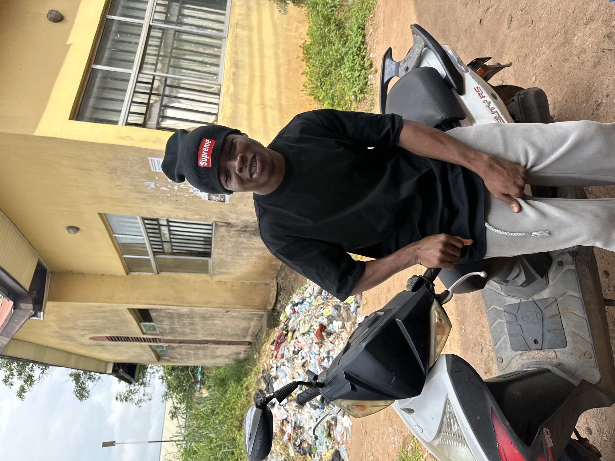 In the city of Ihiagwa, Nigeria, a young Black male wearing a black beanie with a red "Supreme" logo and a black t-shirt, sits on a white scooter. He is also wearing light gray sweatpants with a braided drawstring. The scooter is parked on a dirt road in front of a yellow building with barred windows. To the left of the scooter, a large pile of trash is visible, consisting of various plastic bags and debris. Overcast skies suggest a cloudy day. The scene appears to be an outdoor, possibly residential or commercial, area.
