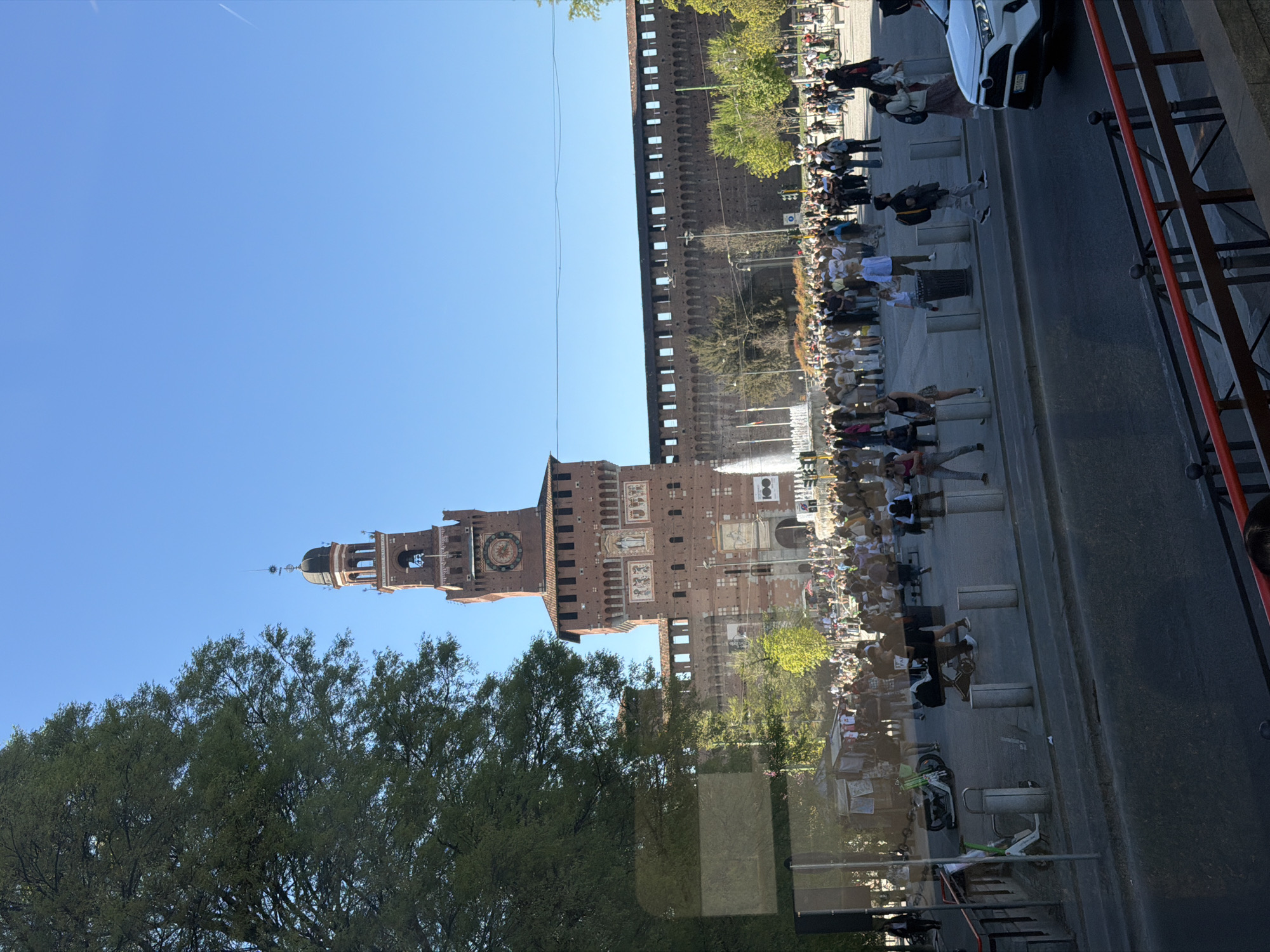 The image shows Castello Sforzesco, a castle in Milan, Italy. The castle, made of red brick, features a tall tower with a clock and a dome, and is surrounded by greenery and a crowd of people. A fountain can be seen in front of the castle. A road runs along the right side of the frame, with a car visible. The sky is a clear blue. The image appears to have been taken from an elevated viewpoint, possibly inside a vehicle.