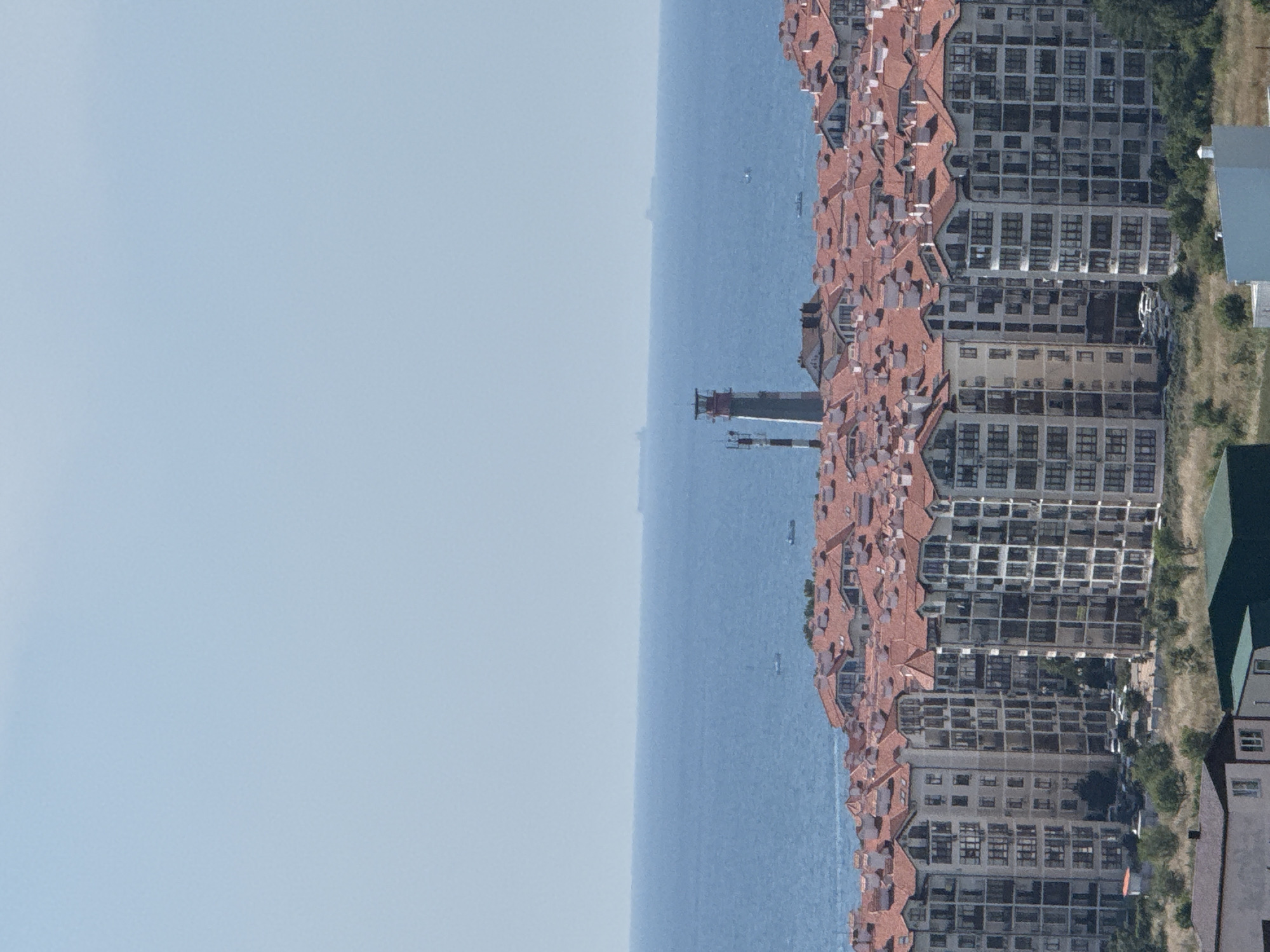 This is a wide shot of a coastal city with many residential buildings. In the foreground, there are several multi-story apartment buildings with red tile roofs. The buildings are densely packed and appear to be modern construction. Some of the buildings have balconies and large windows. In the midground, there is a lighthouse and a body of water that resembles a sea or ocean. There are a few small boats or ships visible on the water. In the background, the sky is clear and blue. The overall impression is of a sunny day in a developed coastal area. This scene is likely from Gelendzhik, Russia, a popular resort city known for its coastline and architecture.