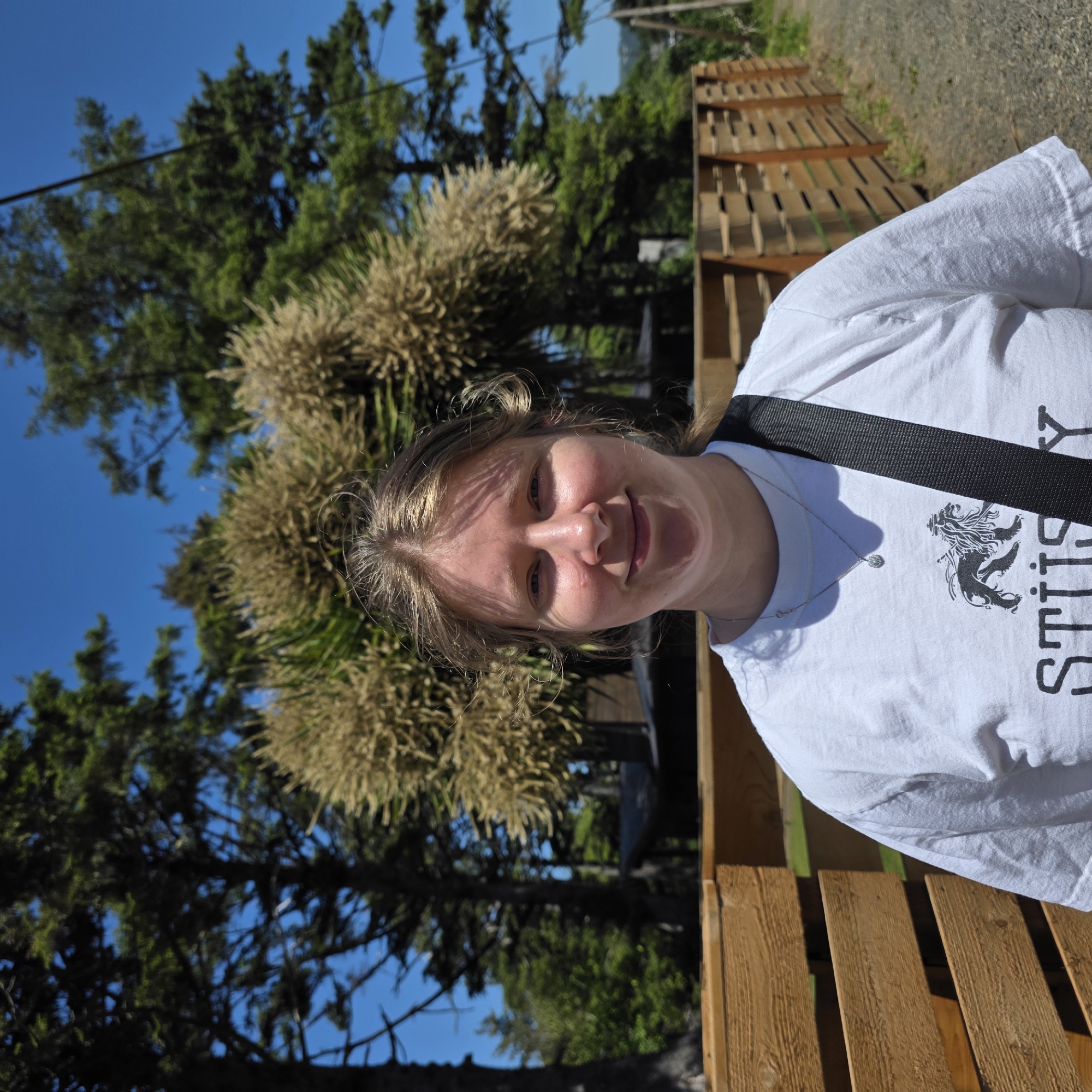 A young woman stands outside in front of a wooden fence and trees. She is wearing a white t-shirt with a black strap over her shoulder. The t-shirt has a graphic of a lion and the word "STUSSY" printed on it. The woman has light skin, light brown hair, and blue eyes. She is smiling and looking at the camera. The background consists of lush green trees and a bright blue sky. The location is Cannon Beach, Oregon, United States of America.