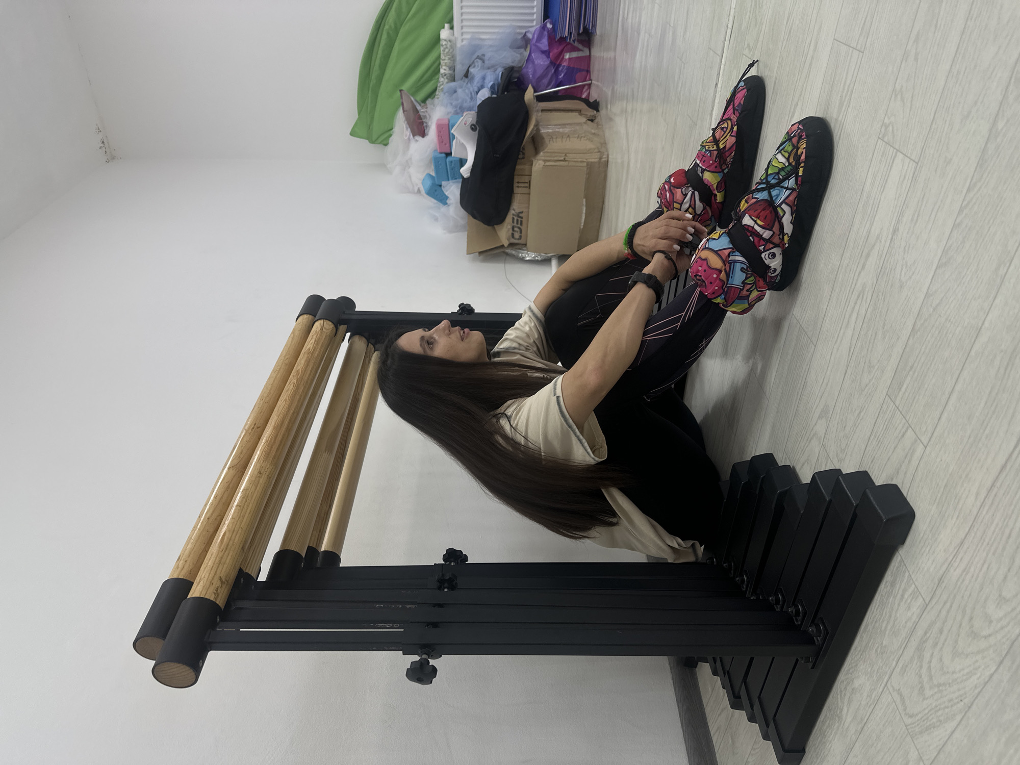 An indoor, side-profile shot shows a woman sitting on the floor next to a piece of exercise equipment. She has long, dark hair and is wearing a light-colored t-shirt over black leggings with pink/purple geometric patterns. Her knees are bent, and she is holding her shins, leaning back against the exercise equipment. She has a watch on her left wrist and green and black bracelets on her right. Her feet are covered in brightly colored, patterned slipper-like footwear, featuring various cartoon designs such as donuts, strawberries, and hearts.
The exercise equipment, likely a ballet barre, is made of black metal framework with several light wooden bars. It appears adjustable, with black knobs visible on the frame. The floor is light wood or laminate.
In the background, behind the woman, there's a cluttered area against a white wall. This area contains stacked cardboard boxes, various plastic bags (one purple), a white radiator, a piece of green fabric (possibly a blanket or towel), and light blue foam blocks. One cardboard box clearly displays the text "СДЭК" (SDEK, a Russian logistics company) and partially visible "ALLA-NA".
The woman's gaze is directed upwards and slightly to her left, with a neutral expression. The setting appears to be a studio or a versatile indoor space in Votkinsk, Russia. The lighting suggests daytime.