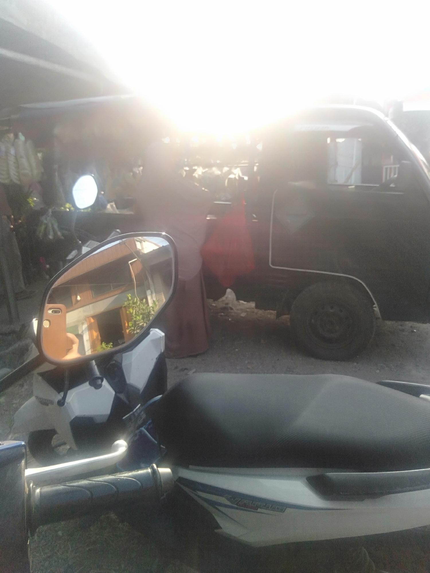 In Sukadami, Indonesia, a side-view mirror of a motorcycle reflects a building with brown siding and green foliage. Behind the motorcycle, a dark-colored pickup truck is parked under a makeshift shelter. The truck has a single rear wheel visible. Sunlight is intensely bright, creating a lens flare across the upper portion of the image. The scene appears to be an outdoor marketplace or roadside area, with various items hanging from the shelter in the background. A person wearing a brown garment is partially visible near the truck.