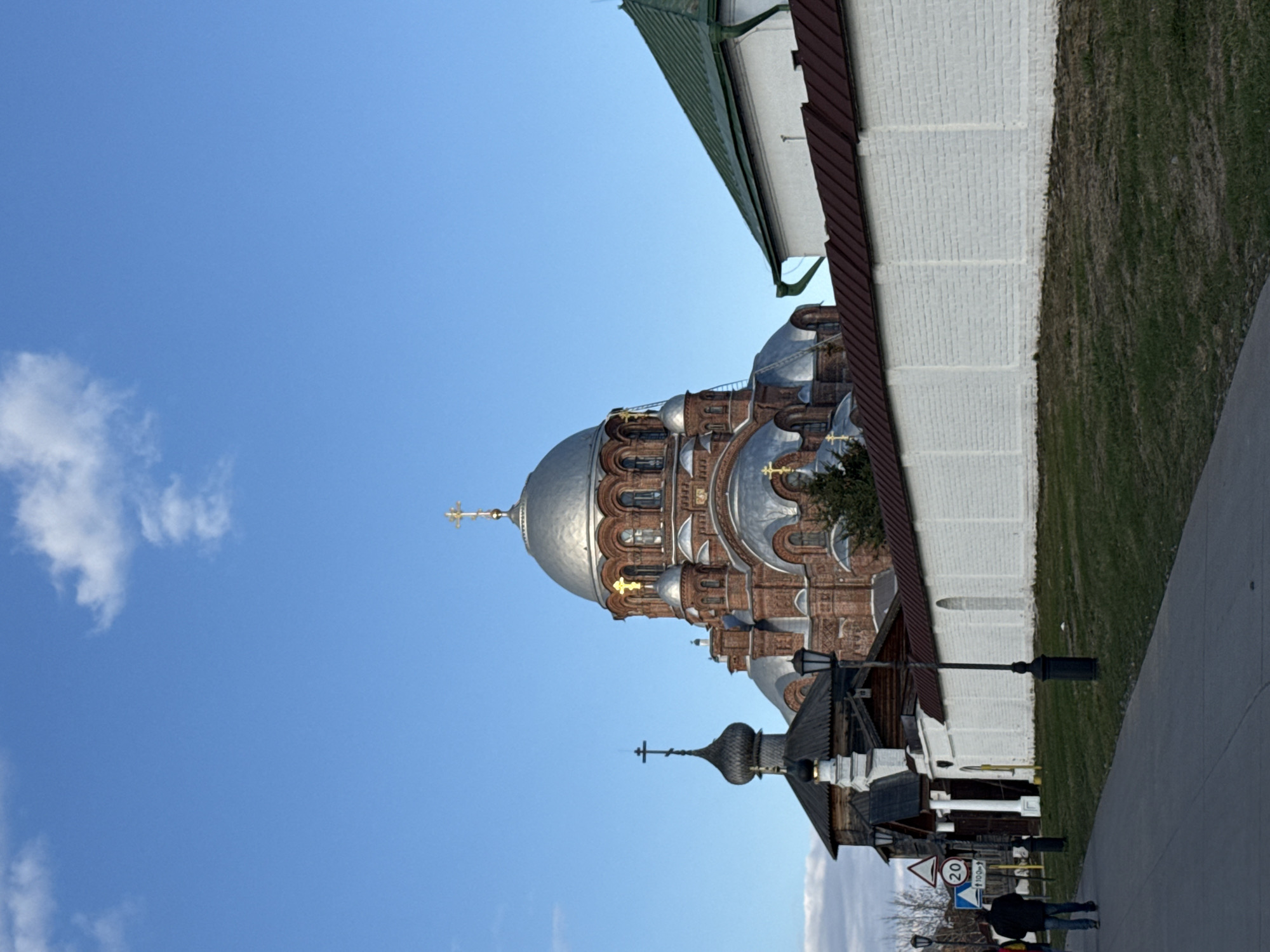 The image shows the Cathedral of the Mother of God of All Who Sorrow in the city of Sviyazhsk, Russia. The cathedral, made of red brick, features silver domes and golden crosses. A white brick wall runs alongside the cathedral, and a small wooden structure with a black dome stands closer to the viewer. A sidewalk is visible in the foreground with two people walking on it. Street lamps and signs are also present. The sky is blue with some clouds.