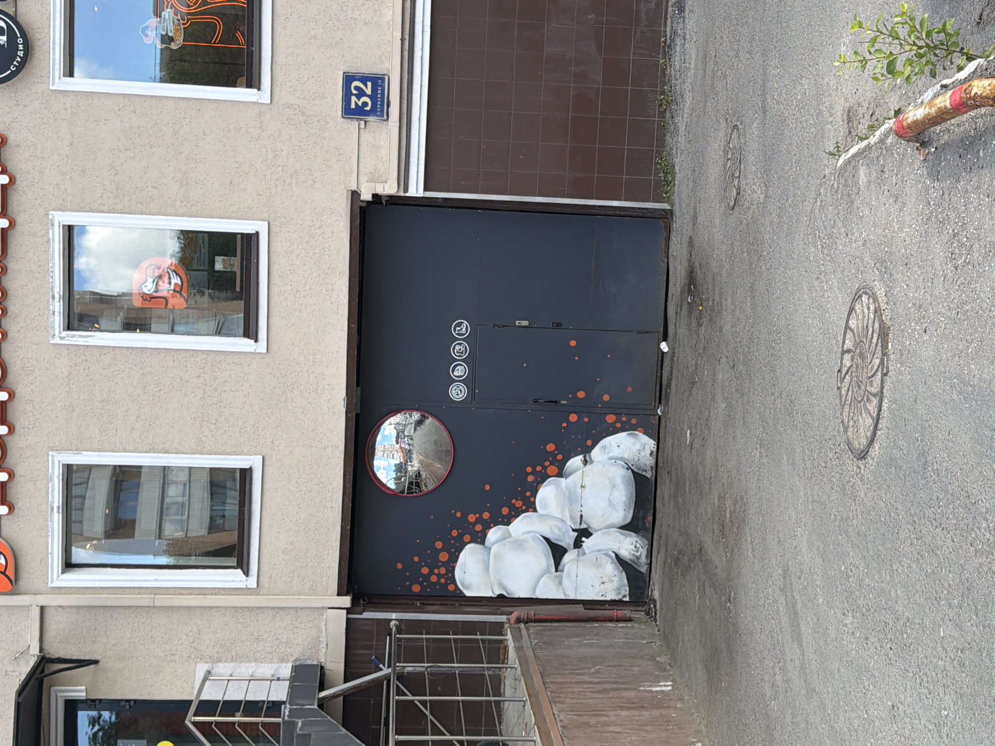 The image depicts the exterior of a commercial building, likely a garage or workshop, in Moscow, Russia. A dark gray, large garage door dominates the center of the frame, featuring a mural of stacked white, amorphous shapes with orange speckles. A convex mirror with a red rim is mounted above the door, reflecting a street scene. To the right of the garage door, a blue sign with the number "32" and the Cyrillic text "СТРОЕНИЕ 13" (Building 13) is affixed to the building's beige facade. Above the garage door, two windows are visible. The left window has a white frame and displays reflections of a building interior, while the right window has a similar frame and shows a reflection of a bright sky and some greenery. The building's facade is textured. In the foreground, a gray asphalt pavement extends towards the viewer, with two circular manhole covers embedded in it. A red and white striped metal barrier and some green foliage are visible on the right side of the pavement, near the building's tiled wall. The overall scene suggests an urban commercial area.