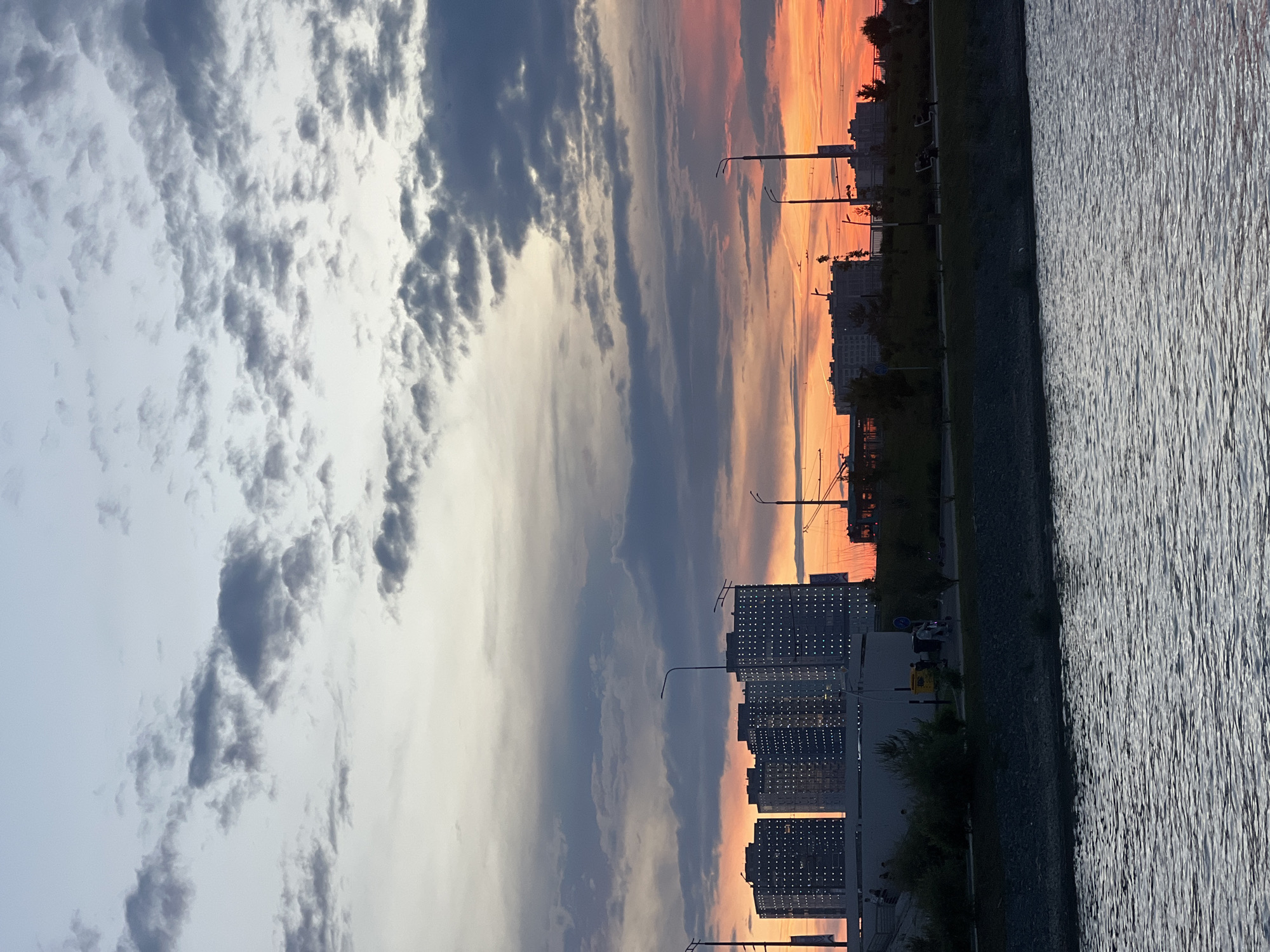 The image captures a sunset over a body of water, likely a river or a bay, in the city of Kazan, Russia. In the distance, a modern cityscape is visible, characterized by several tall residential buildings with numerous illuminated windows. The buildings are silhouetted against a vibrant sky painted with hues of orange, pink, and grey clouds.
Along the embankment, there is a paved walkway with lampposts and scattered trees. A few individuals can be seen walking and cycling on the path. The water in the foreground reflects the sky, creating a shimmering effect. The overall scene suggests an evening urban landscape, tranquil yet alive with the glow of the setting sun and the lit windows of the city.