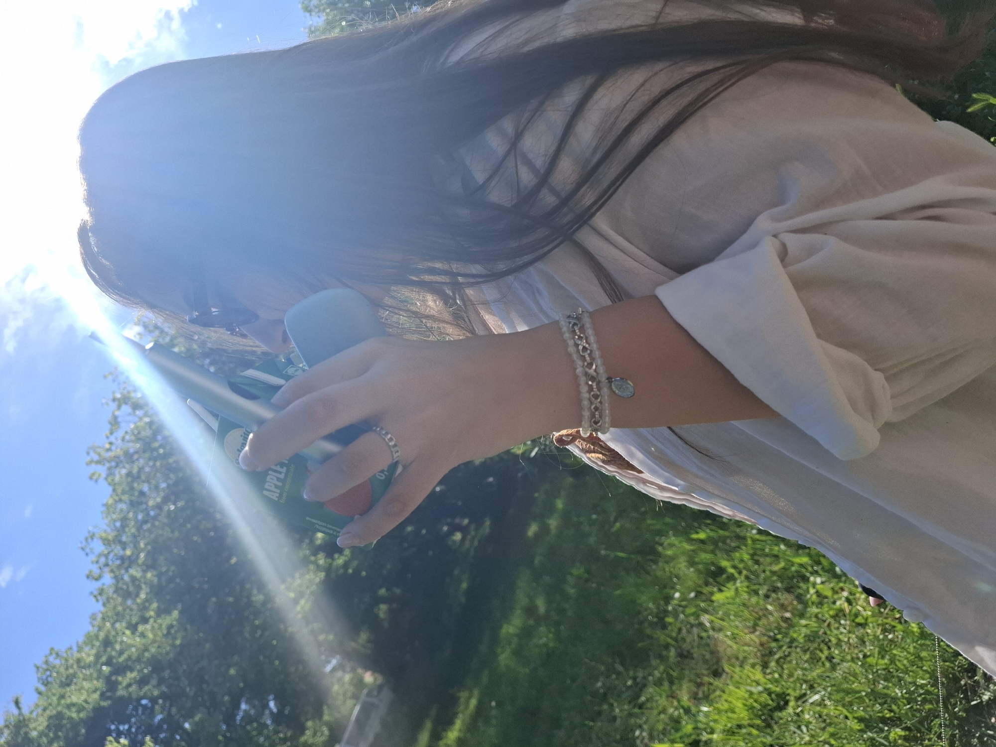 A woman in a white shirt is outdoors on a sunny day, holding an apple juice box in her left hand. She is wearing a silver bracelet and ring. The location is grassy with trees in the background, under a partly cloudy sky. The specific location within Tsarevo, Bulgaria, is not discernible from the image.