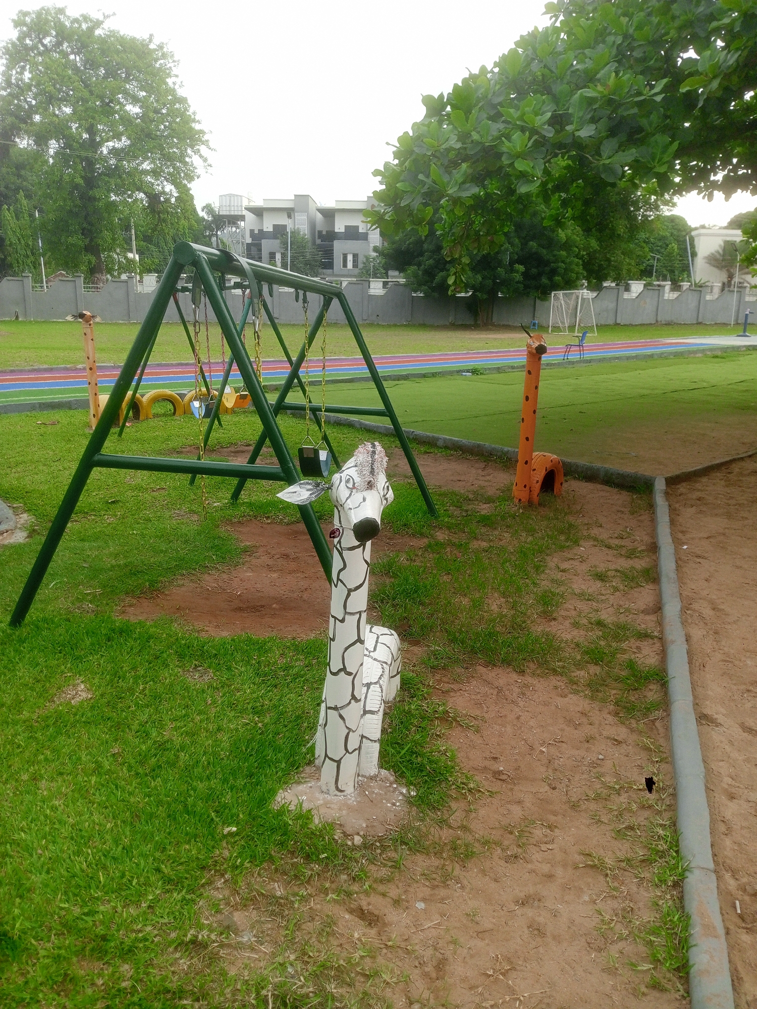 The image depicts an outdoor playground in Abuja, Nigeria. The playground features a green swing set with yellow tire seats and traditional seats. Two giraffe sculptures, one white with black markings and the other orange with black spots, stand on either side of the swing set. A wooden giraffe is in the background. A colorful running track is visible behind the swing set. The ground is a mix of green grass and dirt. A grey concrete curb marks the edge of the play area. A low wall and trees are in the background, with a modern building visible behind the trees. The scene is taken on an overcast day.