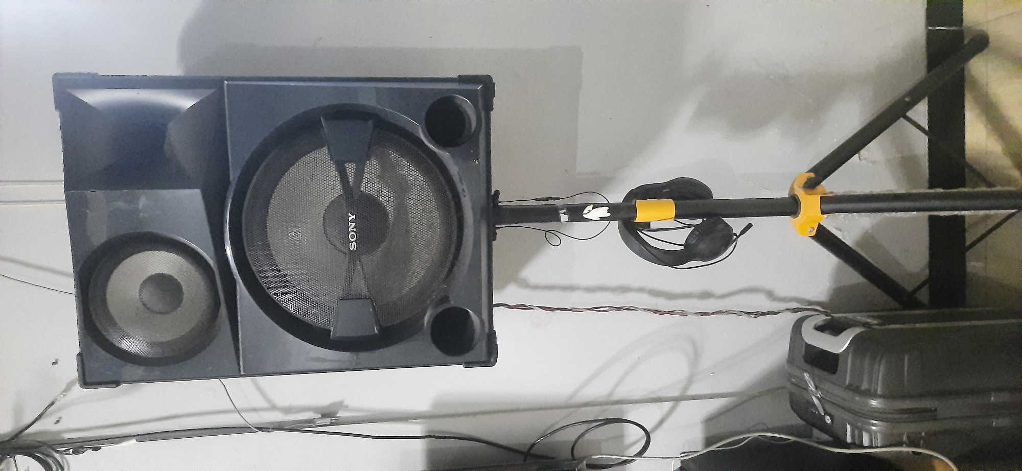A Sony speaker is mounted on a black speaker stand with a yellow accent. A pair of black headphones hangs from the stand. To the left of the speaker stand, a grey suitcase is visible. The background appears to be a plain wall. The scene is indoors, possibly in a residence or storage area.