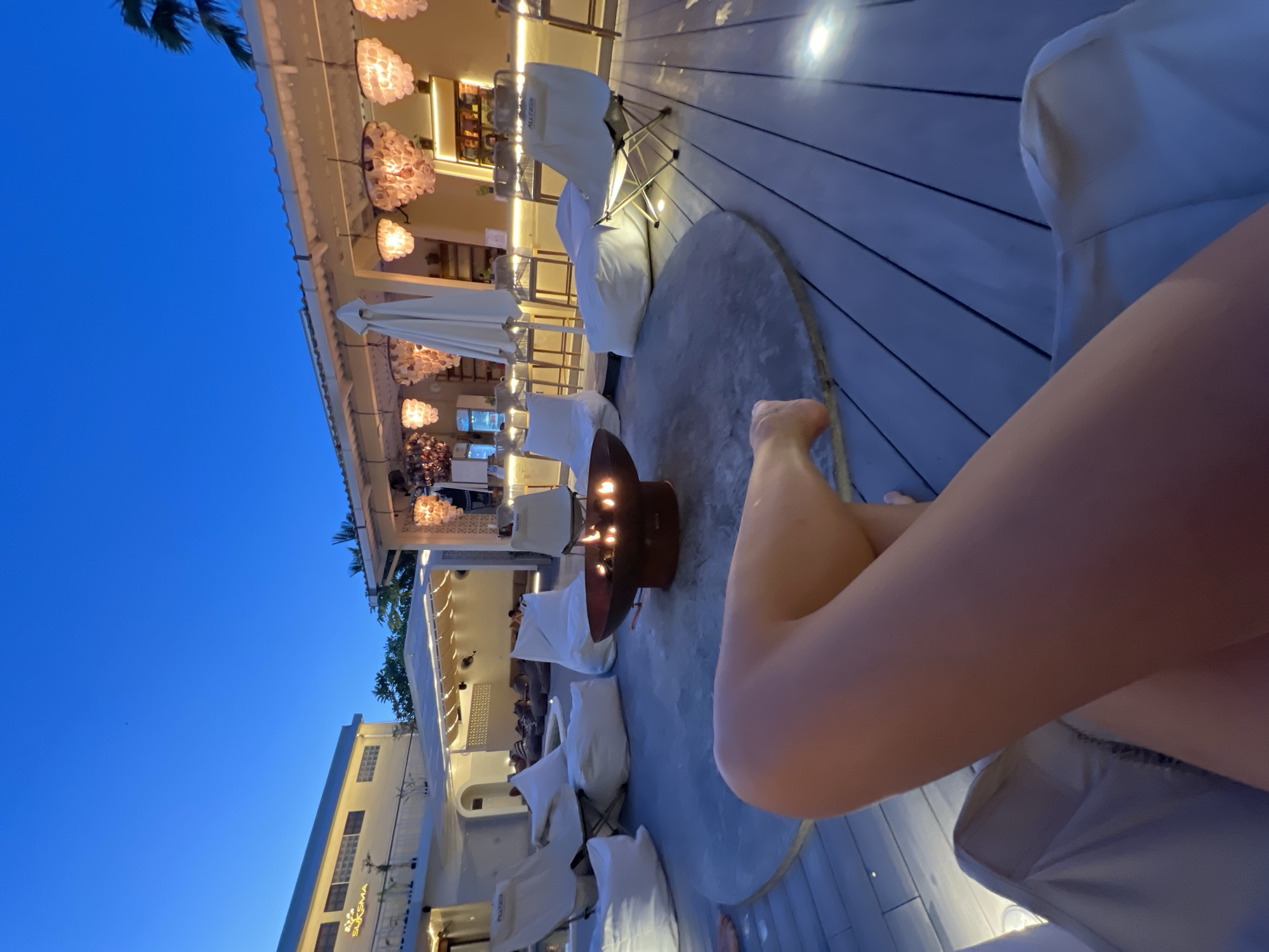 The image depicts an outdoor lounge area at night in North Kuta, Indonesia. A dark blue sky is visible in the upper left corner. The lounge area features a wooden deck with a round, grey rug in the center. A fire pit with burning flames sits on the rug. Several white beanbag chairs and folding chairs surround the fire pit. In the background, a building with multiple lights and white architectural details is visible. A person's legs are prominently featured in the foreground, suggesting they are sitting in the lounge area. The environment is illuminated by the lights from the building and the fire pit, creating a relaxed and inviting atmosphere.