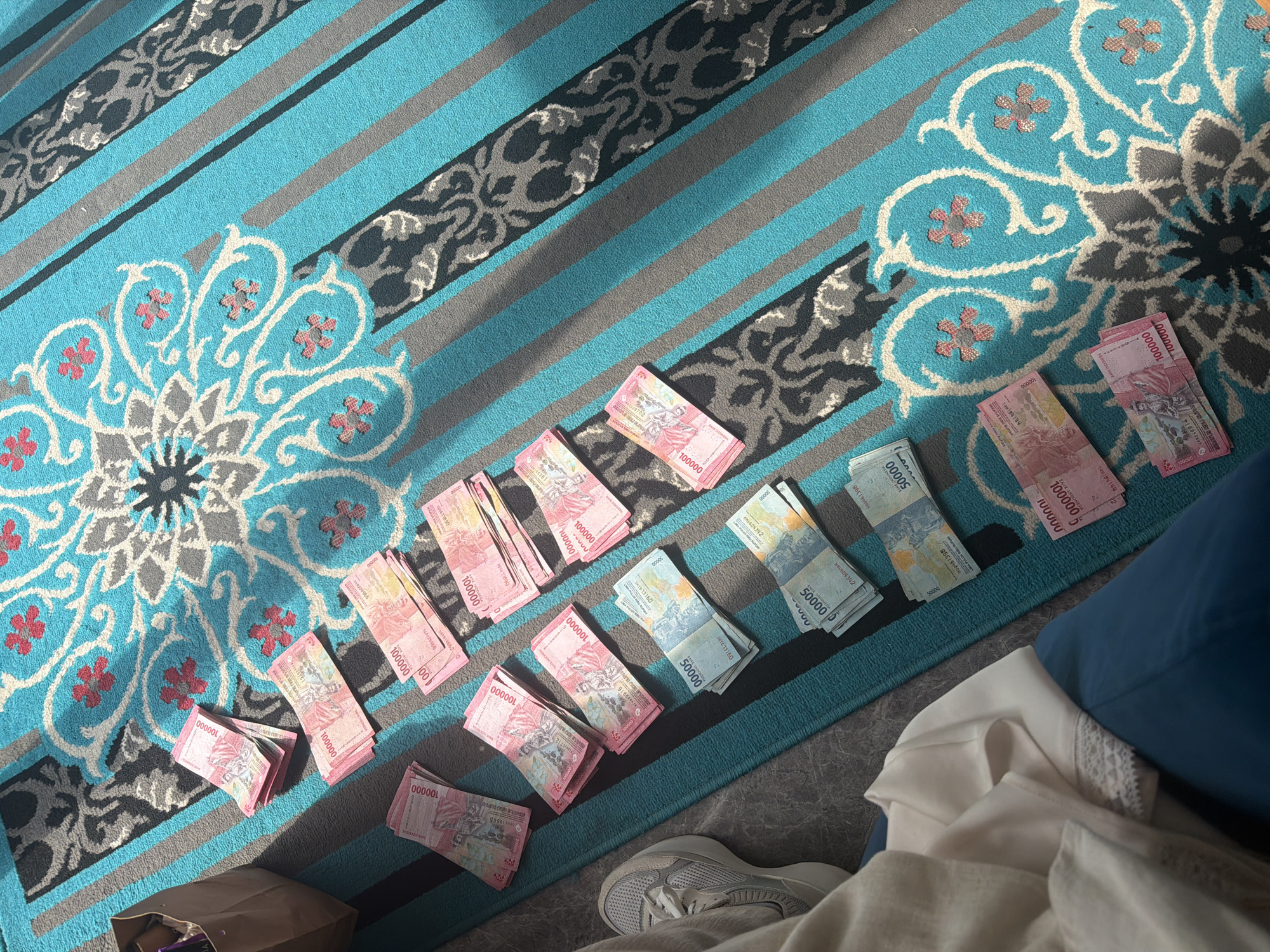 The image shows stacks of Indonesian Rupiah banknotes scattered on a striped and patterned rug. The rug features teal, gray, and black stripes, interspersed with a floral pattern. The banknotes are of 100,000 and 50,000 denominations. Some banknotes are neatly stacked, while others are more loosely arranged. A partially visible paper bag is in the bottom left corner of the image. A gray sneaker, clothing and a blue pillow are also visible. It is not possible to determine the location of Petulu, Indonesia.