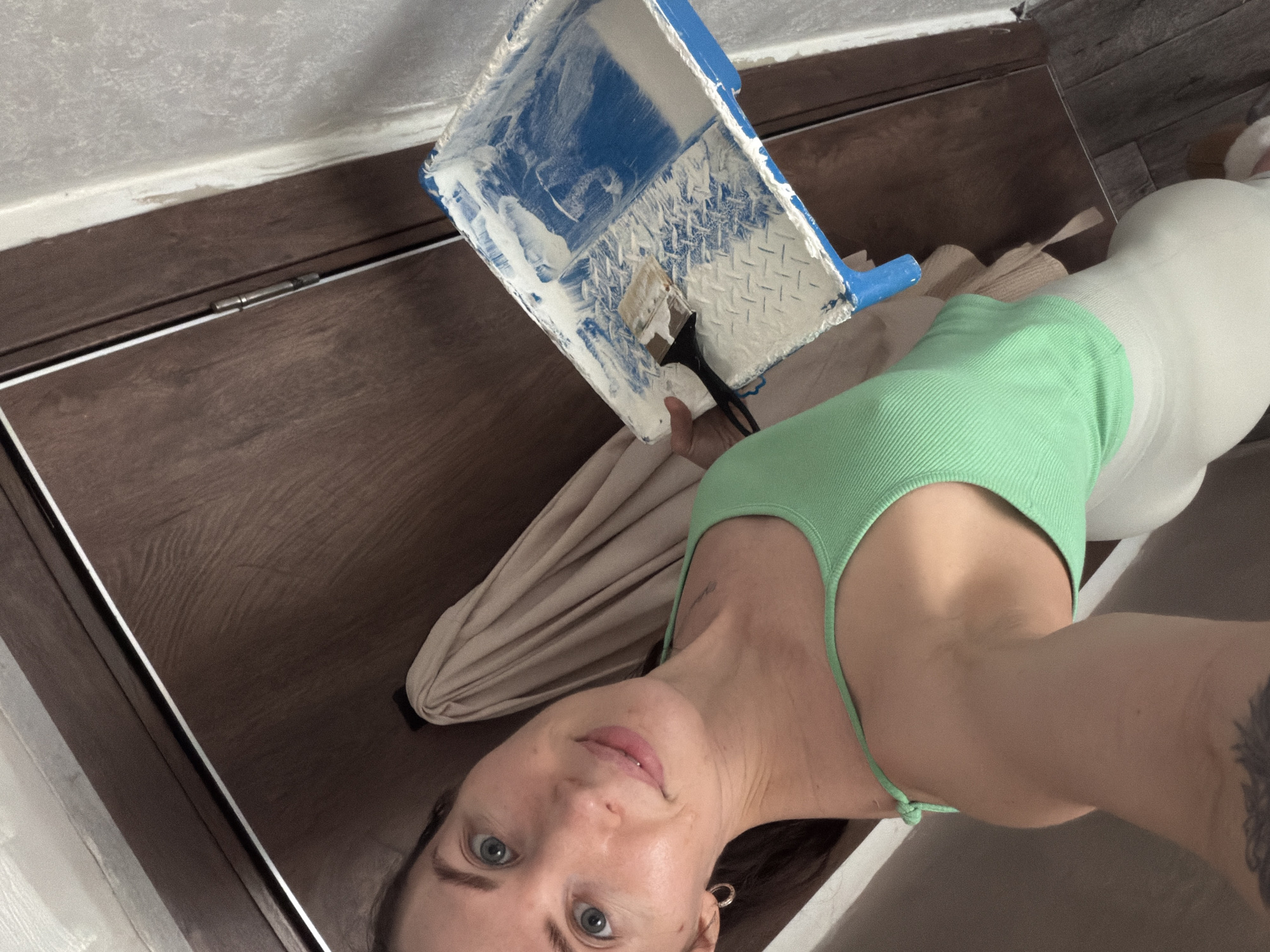 A woman is taking a selfie while painting. She is wearing a green tank top and a light-colored outfit. She holds a blue paint tray with white paint and a brush in it. A drop cloth is also visible. The painting is occurring near a dark wooden surface, likely a baseboard or cabinet. The location may be Perm, Russia.