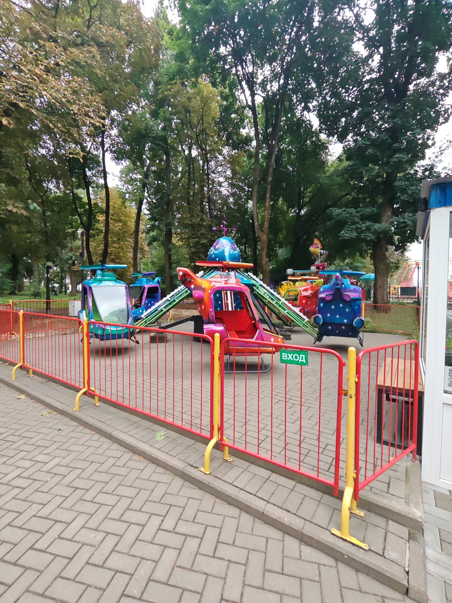 The image depicts a playground ride with multiple helicopter-shaped gondolas situated in an outdoor setting, likely a park. The ride is enclosed by a red metal fence with yellow support posts. The ground is paved with interlocking gray bricks. Several trees with lush green foliage form the backdrop, suggesting it is a warm season. The ride itself is brightly colored, with red, blue, and green dominating the gondolas. A green sign with white Cyrillic lettering, "ВХОД" (which translates to "ENTRANCE"), is visible attached to the fence. In the background, glimpses of other park structures and more trees can be seen. The overall atmosphere is that of a recreational area within a city.