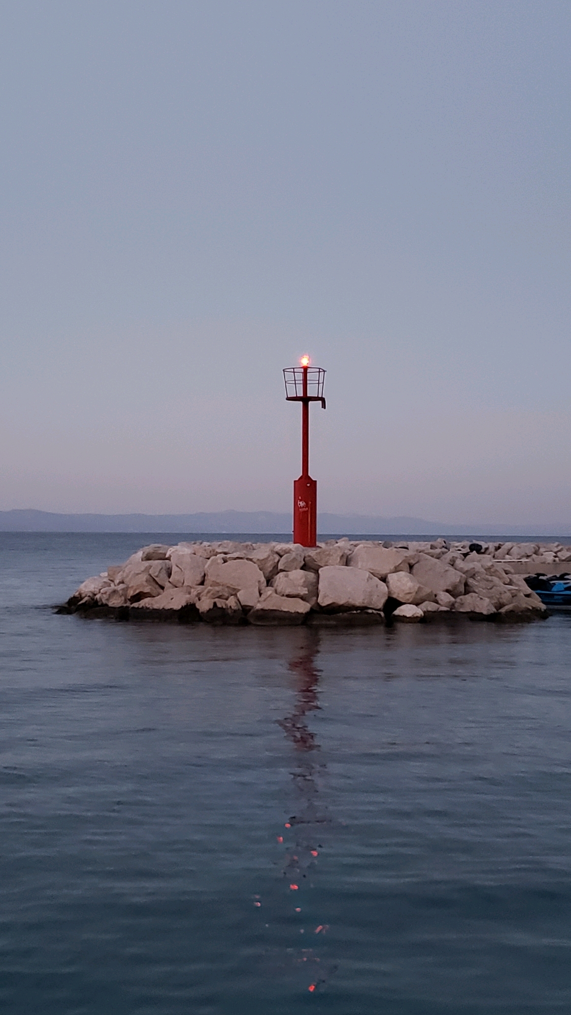 A vertical, eye-level, medium shot shows a red navigational beacon on a man-made breakwater of large, light-colored stones. The beacon is topped with a red light that is illuminated. The breakwater extends into dark blue water, with a hazy, lighter blue sky above, suggesting dawn or dusk. In the distance, a dark, low-lying landmass, likely hills or mountains, is visible. The water reflects the red light of the beacon. A small portion of a blue object, possibly a boat, is visible on the far right of the breakwater. The location is identified as Općina Tučepi, Croatia.