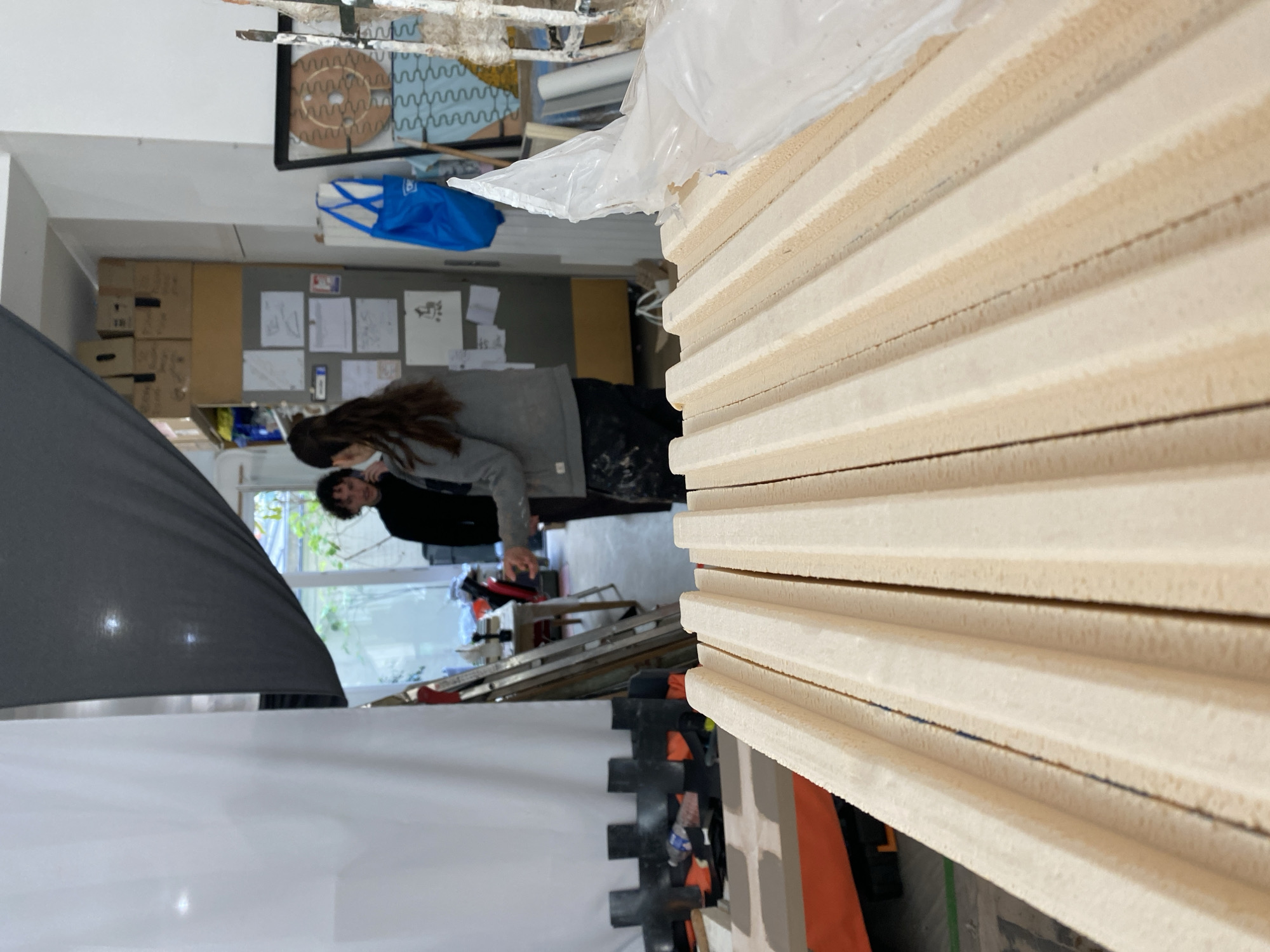 This photograph depicts an interior scene, likely a workshop, studio, or storage area, captured from a slightly elevated perspective. The setting appears busy and functional, filled with various materials and equipment.
In the immediate foreground, dominating the right side of the frame, is a tall stack of light-colored, rectangular panels, possibly made of wood or insulation material. They are neatly layered and partially covered by clear plastic sheeting, suggesting they are new materials ready for use. A faint blue line is visible along the edge of some of these panels.
In the mid-ground, on the left, a large black softbox or light diffuser is prominent, angled towards the left. Next to it, a large white surface or backdrop extends, implying a photography setup or a preparation area. Two individuals are situated further back in the mid-ground. One person, with long dark hair, wears a grey hoodie and appears to be looking down or focusing on an object below. Another person, partially obscured behind the first, wears a dark jacket. Their specific activity is not clear, but they seem engaged in a task or observation within the space.
The background provides more context to the environment. Stacked cardboard boxes line a wall, and a bulletin board or wall section covered with numerous papers and sketches is visible, hinting at creative work or planning. A blue fabric bag hangs in the upper background. Through windows in the distant background, some natural light enters, and glimpses of outdoor greenery are visible, indicating it is daytime. A metal ladder is also present near the individuals. The overall impression is that of an active, practical, or creative workspace. No explicit text is clearly legible in the image.