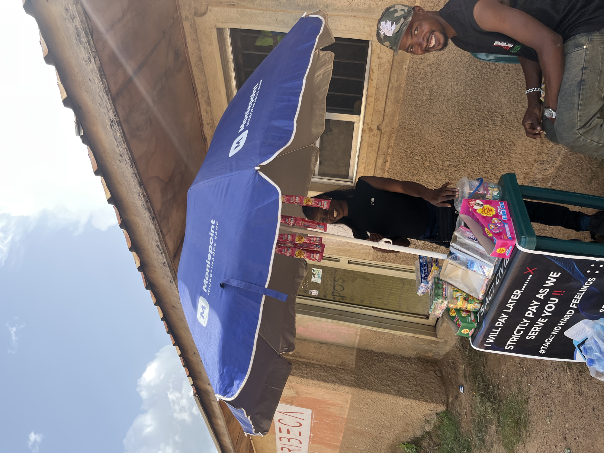 The image depicts an outdoor scene, likely on a street in Jos, Nigeria. A young woman stands behind a small display table laden with various snacks and drinks, including Mentos, Chupa Chups, and bottled water. She is smiling and appears to be attending to customers or waiting for them. A large blue umbrella branded with "Moniepoint Microfinance Bank" provides shade over the display. To the right, a man wearing a camouflage cap and a black t-shirt is seated, also smiling at the camera. A sign on the table reads, "I WILL PAY LATER.......X STRICTLY PAY AS WE SERVE YOU !! #TAG:: NO HARD FEELINGS". The background shows a building with a rough, textured wall and a tiled roof. The overall impression is that of a small roadside business operating in a sunny urban environment.