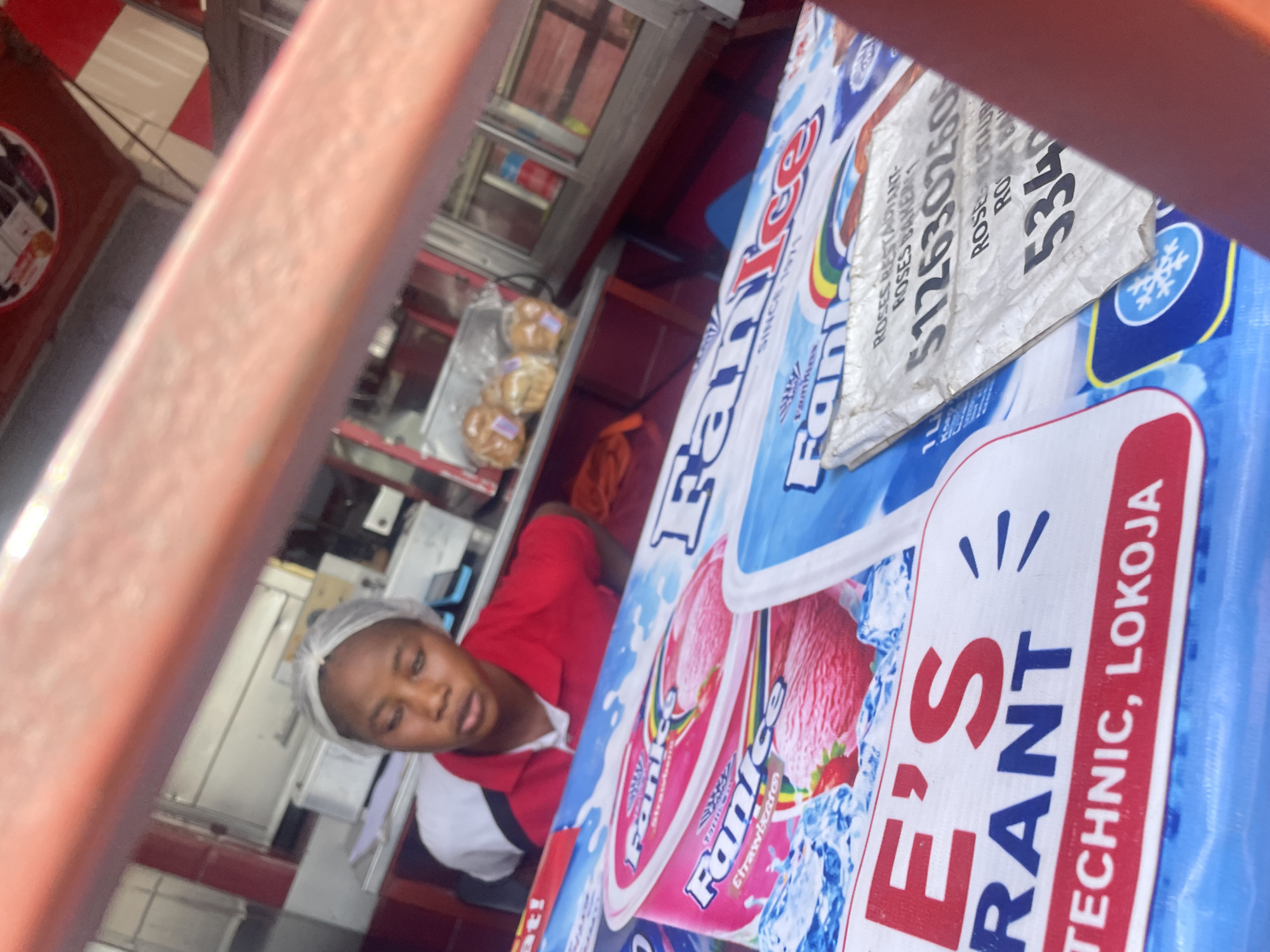 A young woman wearing a hair net and a red and white polo shirt stands behind a counter with ice cream products. The counter is covered with large posters advertising "Fan Ice" and "E's Rant". The "E's Rant" poster has text indicating "TECHNIC, LOKOJA". A small, crumpled white sign with black text is also visible on the counter, displaying "ROSES RESTAURANT- ROSES BAKERY". A phone number "5126302606" and other partial text are on the sign. In the background, baked goods are visible in a display case, and the overall environment suggests a small business or food stall.