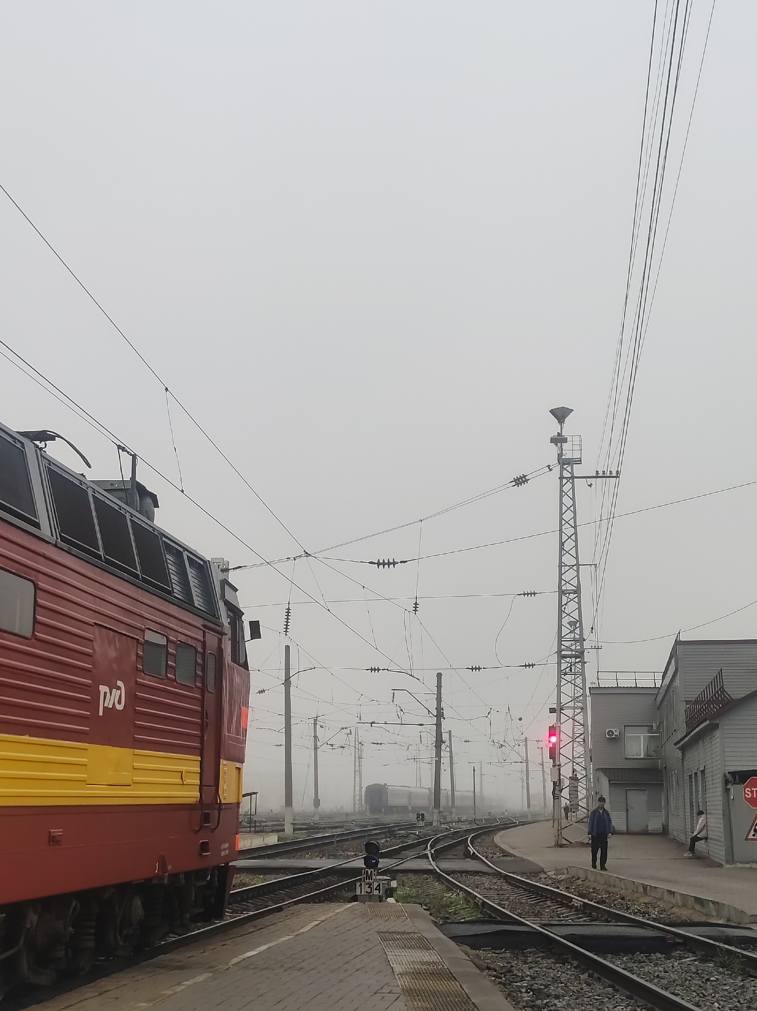 The image depicts a railway station in Kirov, Russia, under an overcast sky. A red and yellow locomotive, identified with the "РЖД" logo, is prominently positioned on the left. The train is positioned on the tracks, indicating it is either stopped or moving slowly. In the background, a signal tower with a red light is visible, suggesting a railway signal in operation. Several power lines and supporting infrastructure are distributed throughout the scene, characteristic of a railway environment. Two individuals are present: one person stands near the signal tower on the platform, and another is seated on a bench outside a building to the right. The building appears to be a station structure, with windows and a covered walkway. The ground is a mix of railway ballast and paved platform sections. The overall atmosphere is muted due to the foggy or overcast conditions.