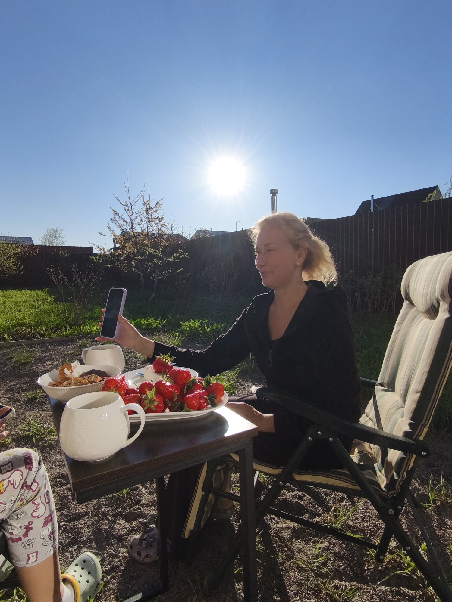 The image shows a woman seated at a small, dark-topped table outdoors in what appears to be a backyard or garden setting, consistent with the location in Steklyannyi, Russia. A clear blue sky with bright sunlight is visible.
The woman, who has blonde hair pulled back, is wearing a dark zippered jacket. She is holding a mobile phone in her left hand and appears to be taking a photo or video. On the table, there are two white serving dishes, one filled with pastries and the other piled high with fresh strawberries. Two large white mugs also sit on the table.
A striped folding chair is positioned to the woman's right. To the left of the table, a person's legs are visible, wearing patterned pajamas and green Crocs. The ground is covered with dirt and sparse grass. In the background is a wooden fence and some foliage. The overall setting suggests a casual, outdoor meal or gathering.