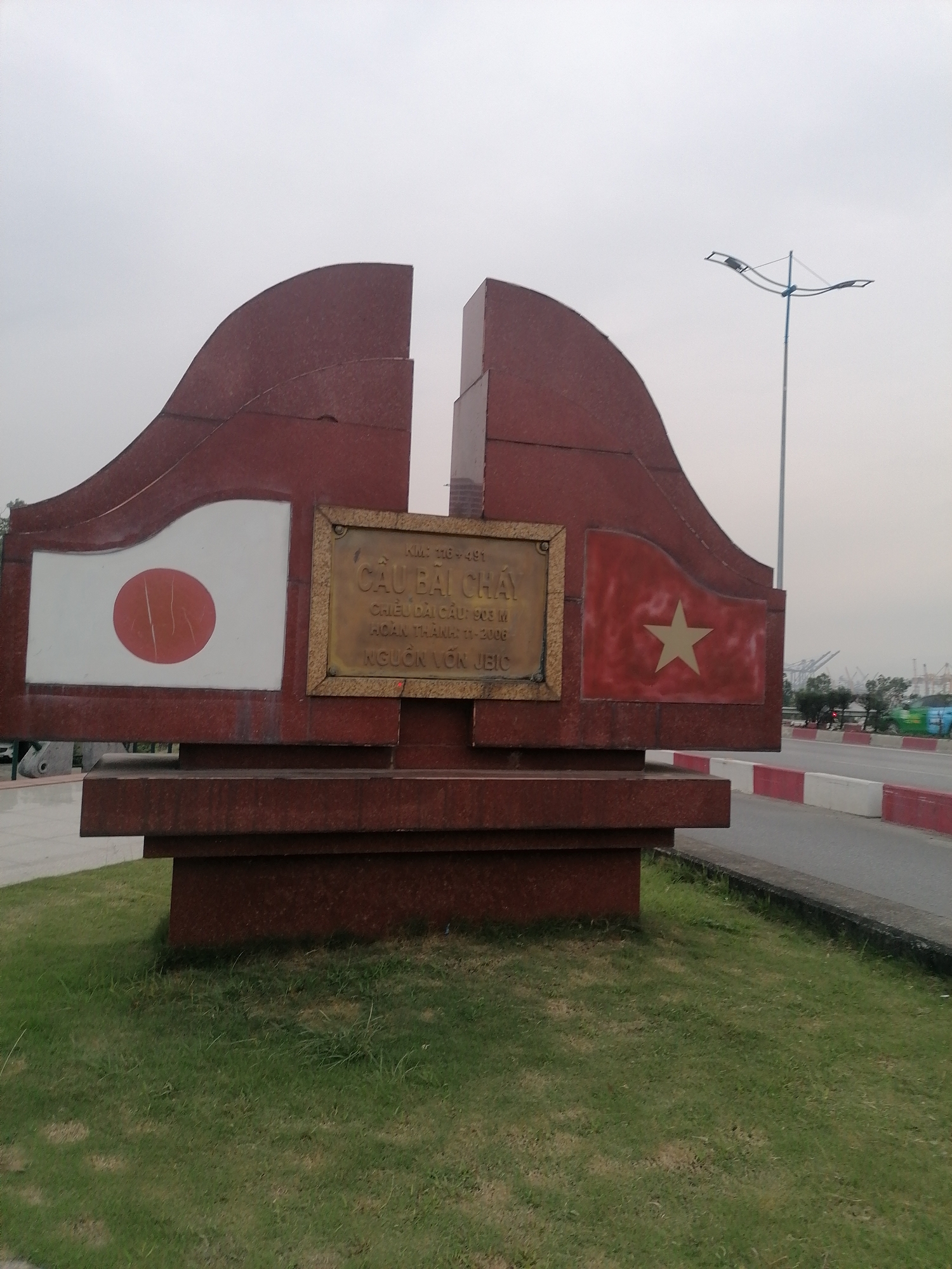 A large, reddish-brown monument stands prominently on a patch of green grass beside a road, under a cloudy, overcast sky. The monument has a distinctive, abstract shape, appearing as two stylized, curved structures that mirror each other, separated by a central vertical gap.
On the left side of the monument, a depiction of the Japanese flag (a red circle on a white rectangle) is visible. On the right side, the national flag of Vietnam (a yellow star on a red rectangle) is displayed.
Centered between these two flags, a golden rectangular plaque is affixed to the monument. The text on the plaque, in Vietnamese, reads: "KM: 110-491", "CẦU BÃI CHÁY" (Bai Chay Bridge), "CHIỀU DÀI CẦU 903 M" (Bridge length 903 m), "HOÀN THÀNH: 11-2006" (Completed: November 2006), and "NGUỒN VỐN JEIC" (Funded by JEIC).
To the right of the monument, a paved road with a distinctive red and white painted curb is visible. A tall streetlight with multiple lamps extends upwards from the right side, silhouetted against the bright, hazy sky. In the distant background, industrial structures, possibly large cranes associated with a port or shipyard, can be seen under the diffused light, hinting at the location's proximity to a coastal or riverine industrial area in Bai Chay Ward, Vietnam. The overall impression is one of a commemorative marker for a significant infrastructure project. No people are present in the frame.