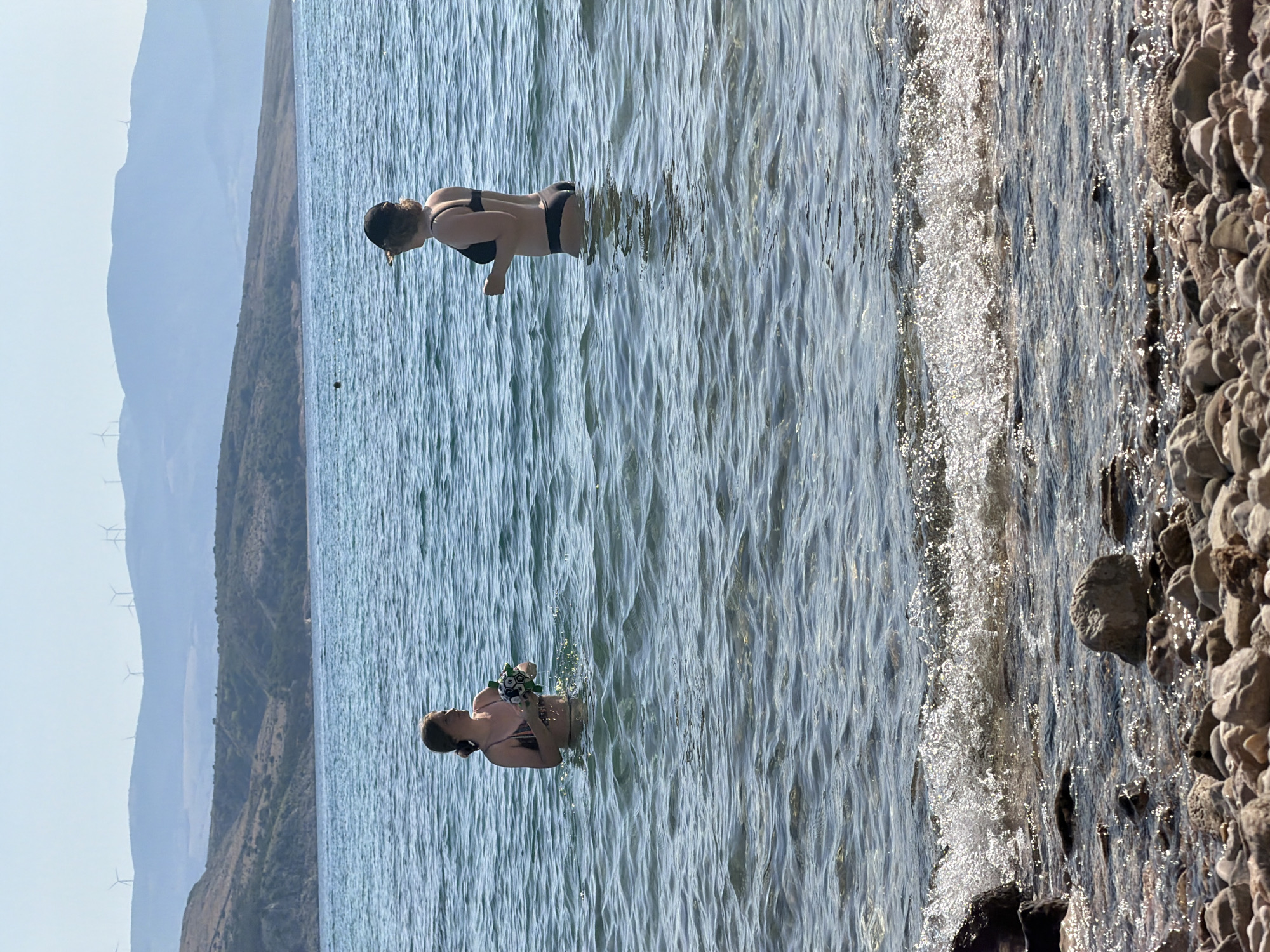 Two individuals are standing in the shallow water of a sea, facing away from the viewer. The person on the left is a woman in a patterned bikini, holding a multi-colored ball. The person on the right is also a woman, wearing a black bikini and a dark baseball cap. In the background, a coastline with hills and scattered wind turbines is visible under a clear sky. The foreground shows a pebbled shore with small waves breaking. The scene appears to be a natural coastal area in Općina Baška, Croatia.