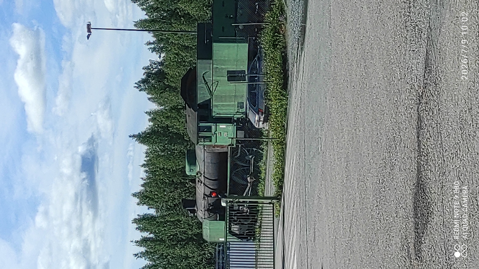 A weathered, green steam locomotive with the number "1011" visible on its side is parked on a railway track in Otanmäki, Finland. The locomotive is partially obscured by a metal fence in the foreground. A dark-colored car is parked behind the fence, near the locomotive. The scene is set against a backdrop of dense green trees and a sky with scattered white clouds. The overall impression is one of a historical or abandoned site.