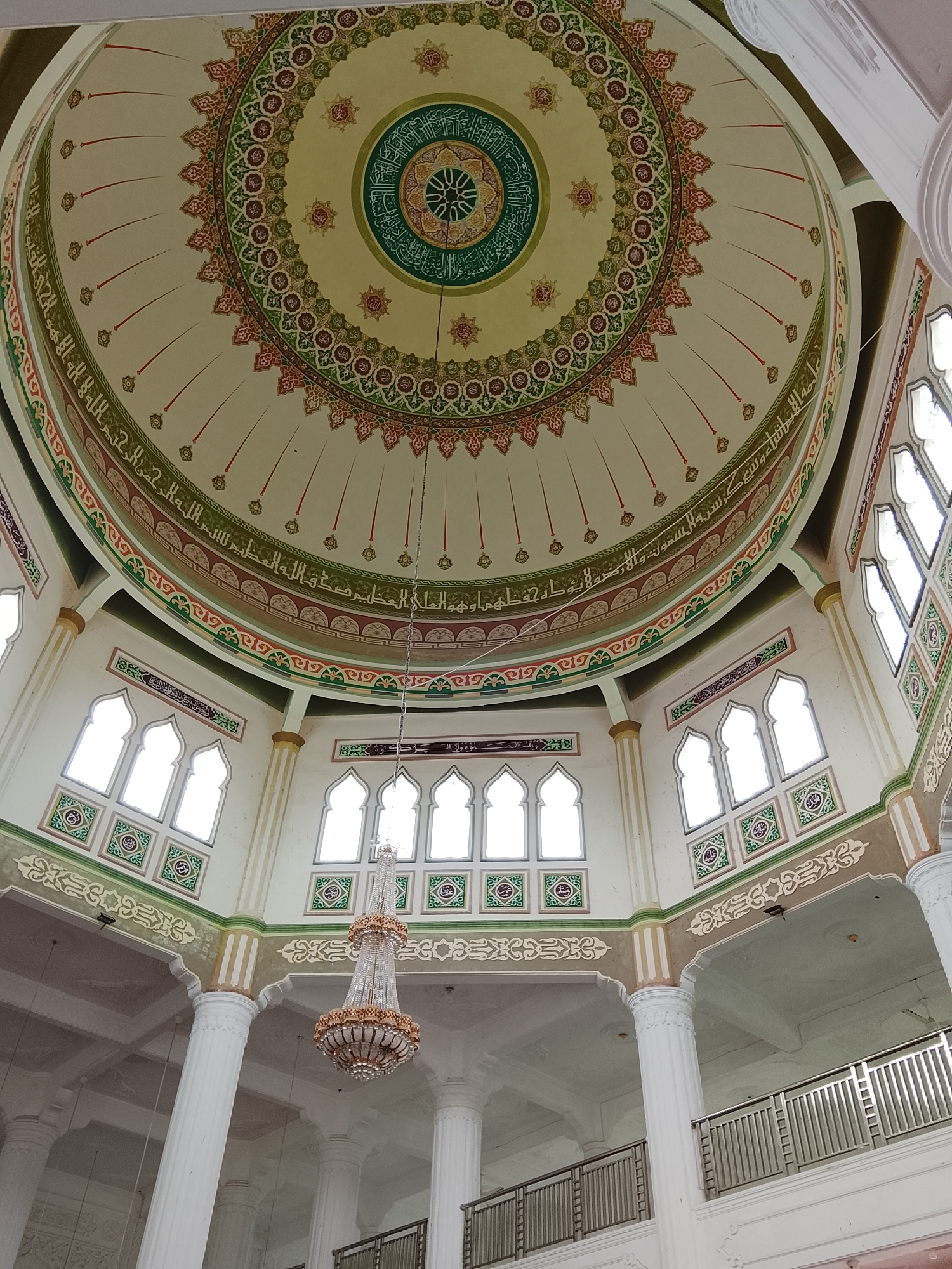 The image shows the interior of a mosque with a domed ceiling and ornate decorations. The dome features intricate Arabic calligraphy and geometric patterns in green, red, and gold. Below the dome, there are arched windows with stained glass or decorative panels. Large white pillars support the structure, and a crystal chandelier hangs from the ceiling. Balconies with metal railings are visible on upper levels. The mosque is located in Lhokseumawe, Indonesia. No people are visible in the image.