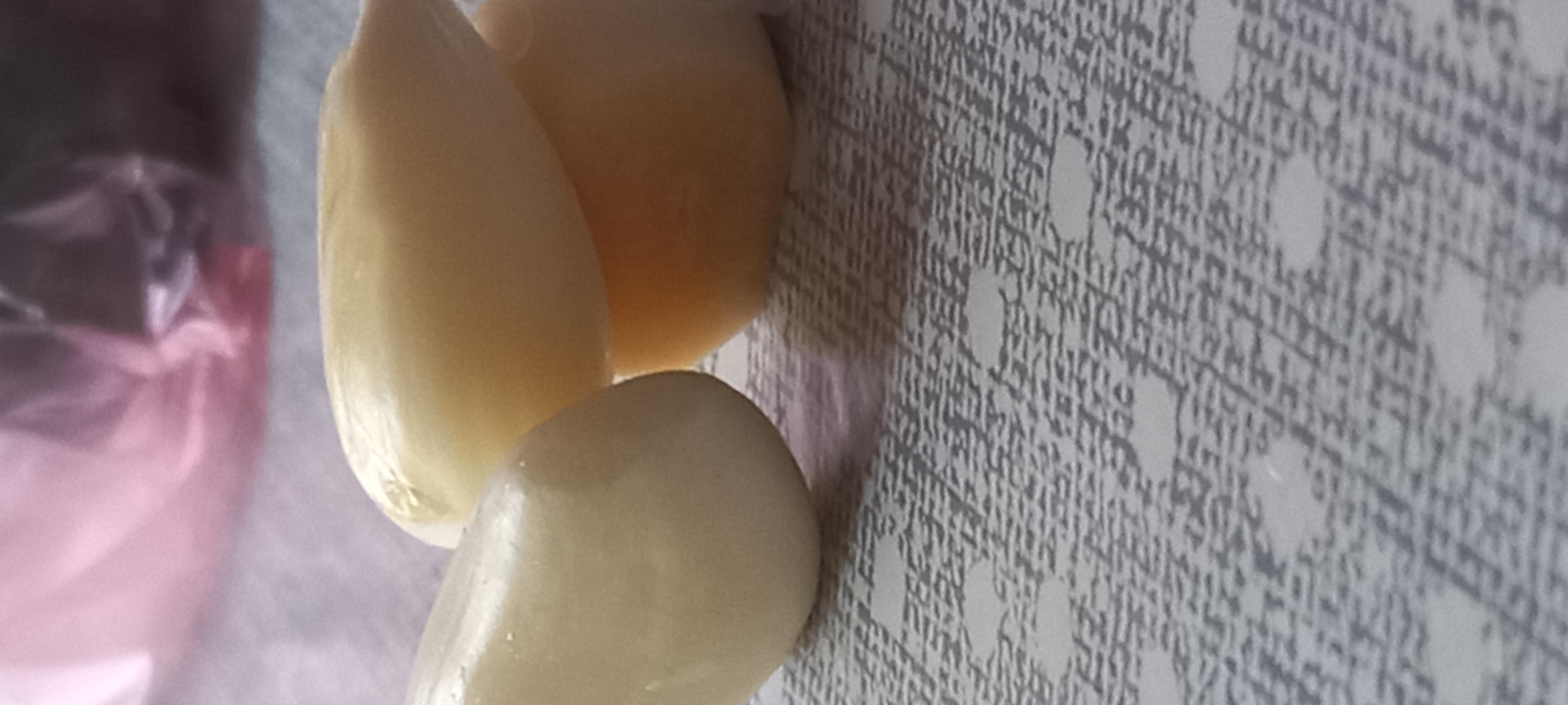 The image displays three peeled garlic cloves positioned on a textured surface. One elongated garlic clove is prominently featured on the left, while two other peeled garlic cloves are situated to the right, appearing to be partially stacked. All three cloves are light cream to pale yellow in color, exhibiting a smooth, moist surface. They rest on a gray surface with a repeating pattern of darker gray grid lines and scattered lighter, irregular circular or oval shapes, suggesting a fabric placemat or textured tabletop. The background is heavily blurred and out of focus, showing indistinct forms and colors, specifically a soft pink or purple hue on the upper left, possibly from a plastic bag or fabric, and darker, less defined shapes on the upper right. The lighting is soft and ambient. The scene is located in the city of Kryvyi Rih, Ukraine.