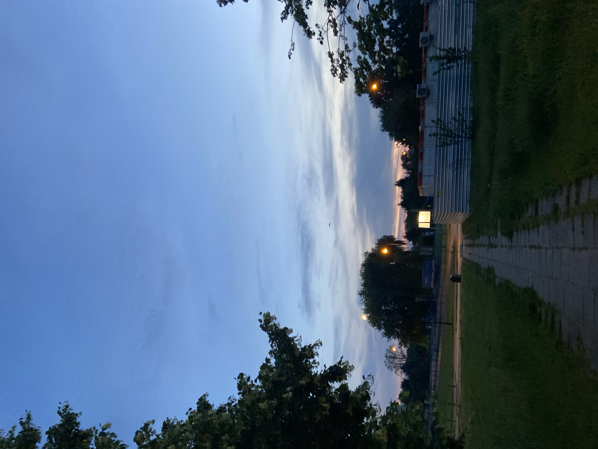The image captures a late evening scene in a city park in Warsaw, Poland. The sky is a gradient of deep blue at the top, transitioning to lighter blues and hints of pink and orange near the horizon, suggesting either sunrise or sunset. Several wispy clouds are scattered across the sky.
In the foreground, a paved walkway made of concrete slabs runs through a grassy area. Tall trees with dense foliage frame the left and right sides of the image, with their branches extending into the sky.
In the middle ground, there are more trees and streetlights. The light from at least two streetlights is visible, casting a warm glow. Behind the trees and lights, there's a construction site enclosed by a corrugated metal fence. The fence has some visible air conditioning units mounted on top of a structure behind it, and a bright white light is emanating from a sign or fixture on the left side of the construction area.
The overall impression is a quiet urban environment during twilight, possibly a park or a recreational area adjacent to a construction zone. No people are visible in the image.