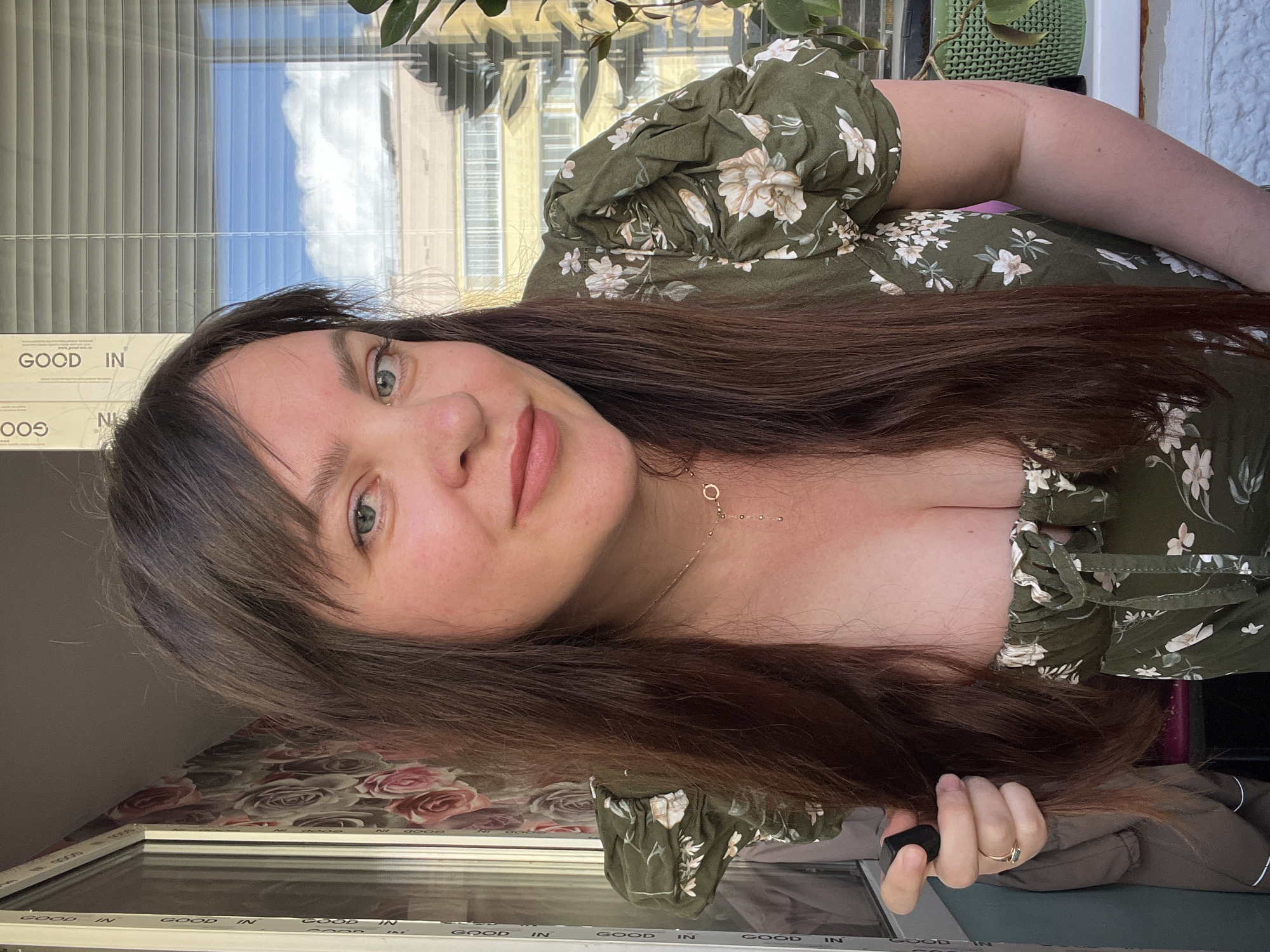 This is a selfie of a young woman with long, dark brown hair, taken indoors. She is wearing a green, floral-print top with puffy sleeves. The woman is looking at the camera with a slight smile and her head is tilted. Her makeup includes pink lipstick and mascara. She is wearing a delicate gold necklace with small circular charms.
The background shows a window with Venetian blinds, through which a blue sky with white clouds is visible, suggesting it is daytime. There are also some plants visible, one in a green patterned pot. Below the window is a patterned wallpaper with roses in shades of pink and grey, and a white window frame with "GOOD IN" printed repeatedly along it. The overall impression is that of a casual, personal moment captured at home.