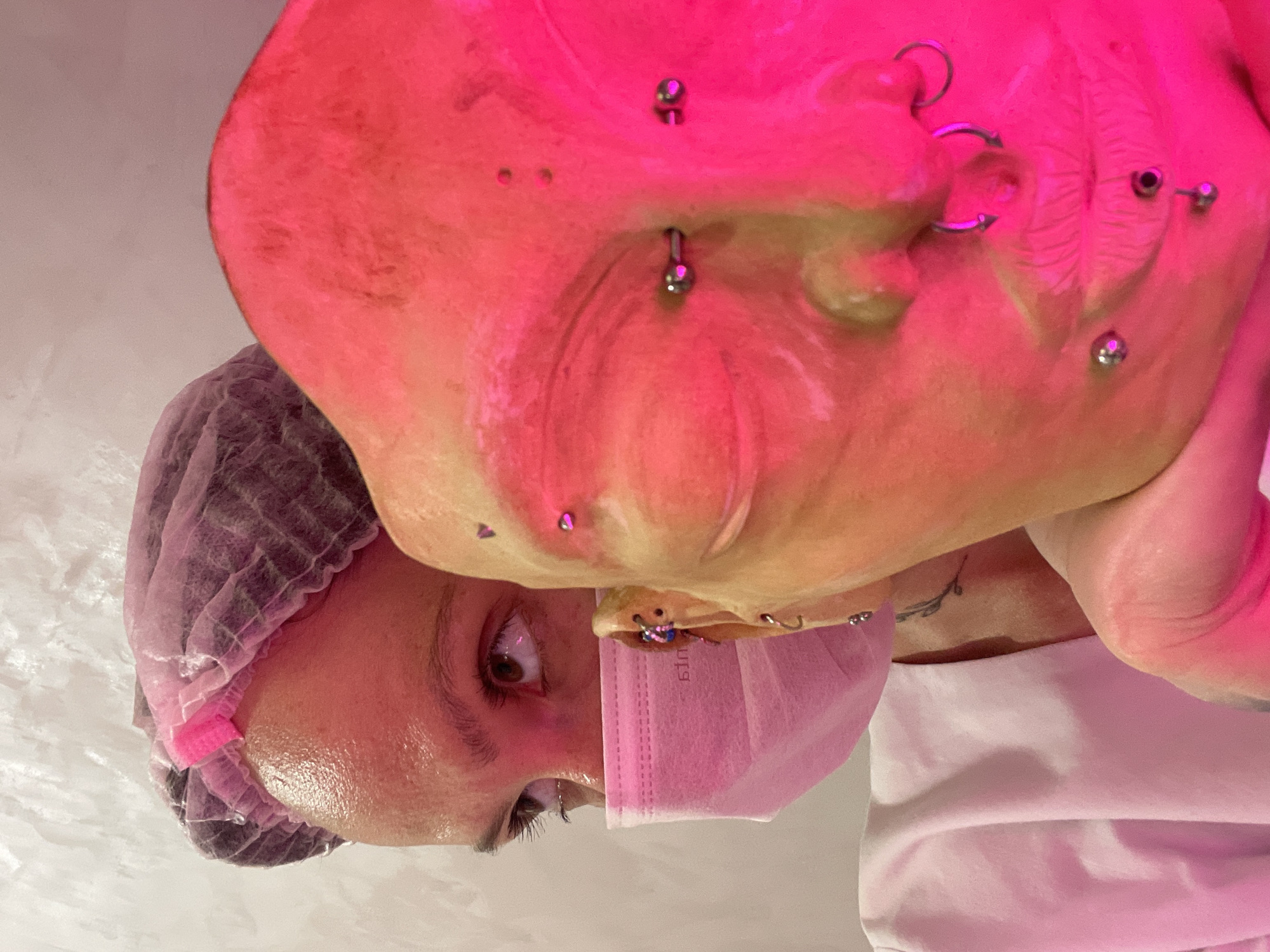 A woman wearing a pink surgical mask and hairnet lies on her back. Her face has multiple piercings. A portion of the room is visible, a sterile, white background. The city is not discernible.