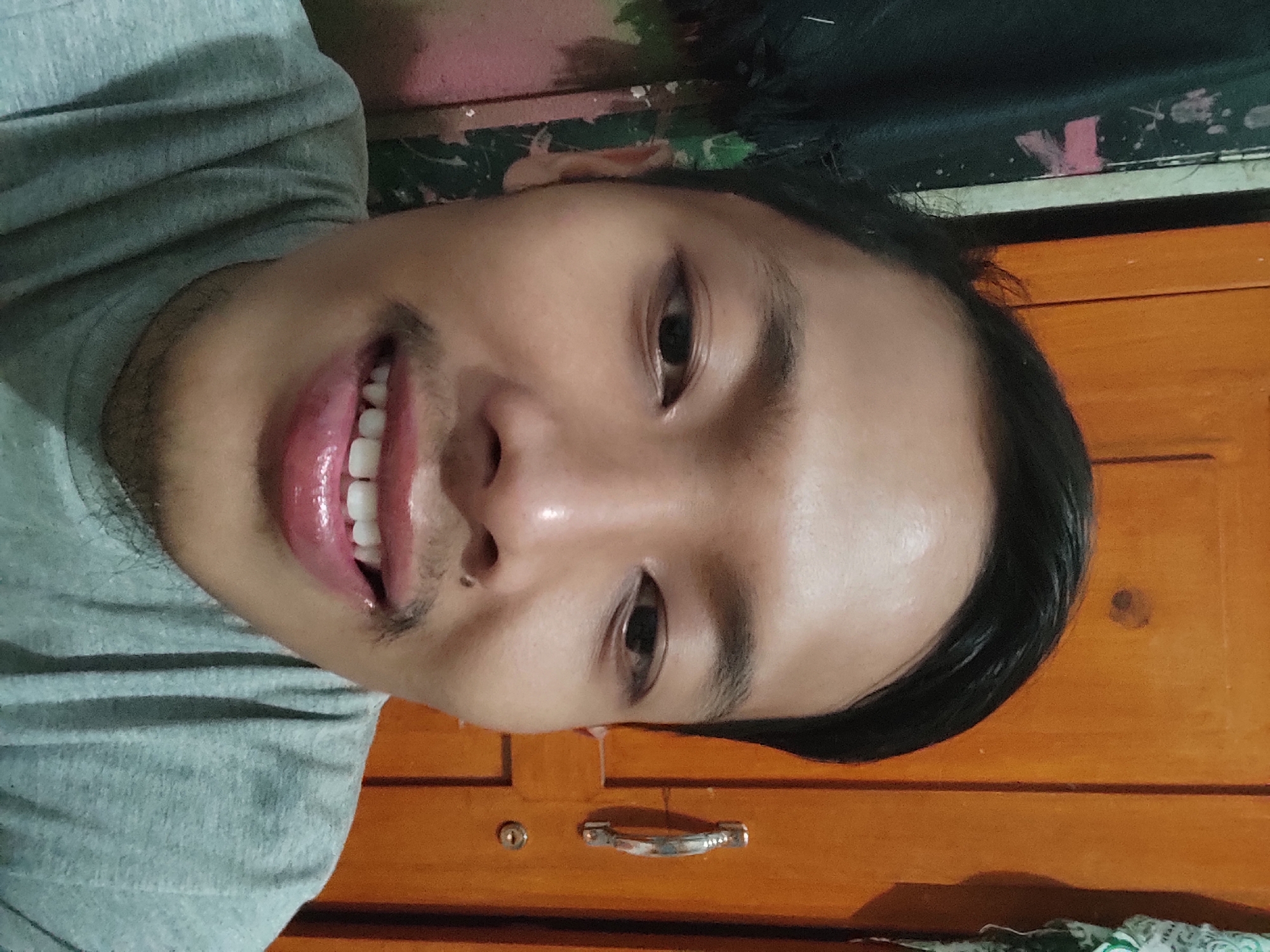 A close-up, waist-up selfie of a young adult male with fair skin, dark hair, and a mustache. He is wearing a gray t-shirt and smiling, showing his teeth. He has a small mole on his upper lip. The background appears to be indoors, with a wooden cabinet to the right and a dark, possibly fabric-covered surface to the left. The lighting is direct and casts a slight sheen on the subject's face. The image was taken in the city of Sumedang, Indonesia.