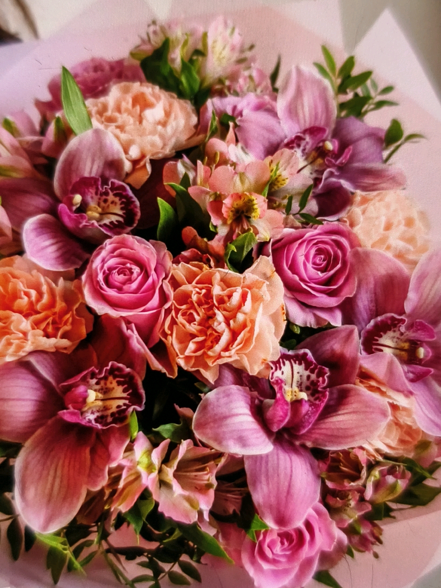 A close-up, top-down, angled shot shows a bouquet of flowers, primarily in shades of pink, purple, and peach. Prominent flowers include orchids, roses, and carnations, interspersed with delicate, star-shaped pink flowers and green leaves. The flowers are arranged tightly, with some petals overlapping. The background is a soft pink, suggesting the bouquet is wrapped in paper or placed against a similar colored surface. The lighting creates soft shadows, highlighting the textures and depth of the floral arrangement. The image does not provide any information about a specific location in Umm Al Quwain, United Arab Emirates.