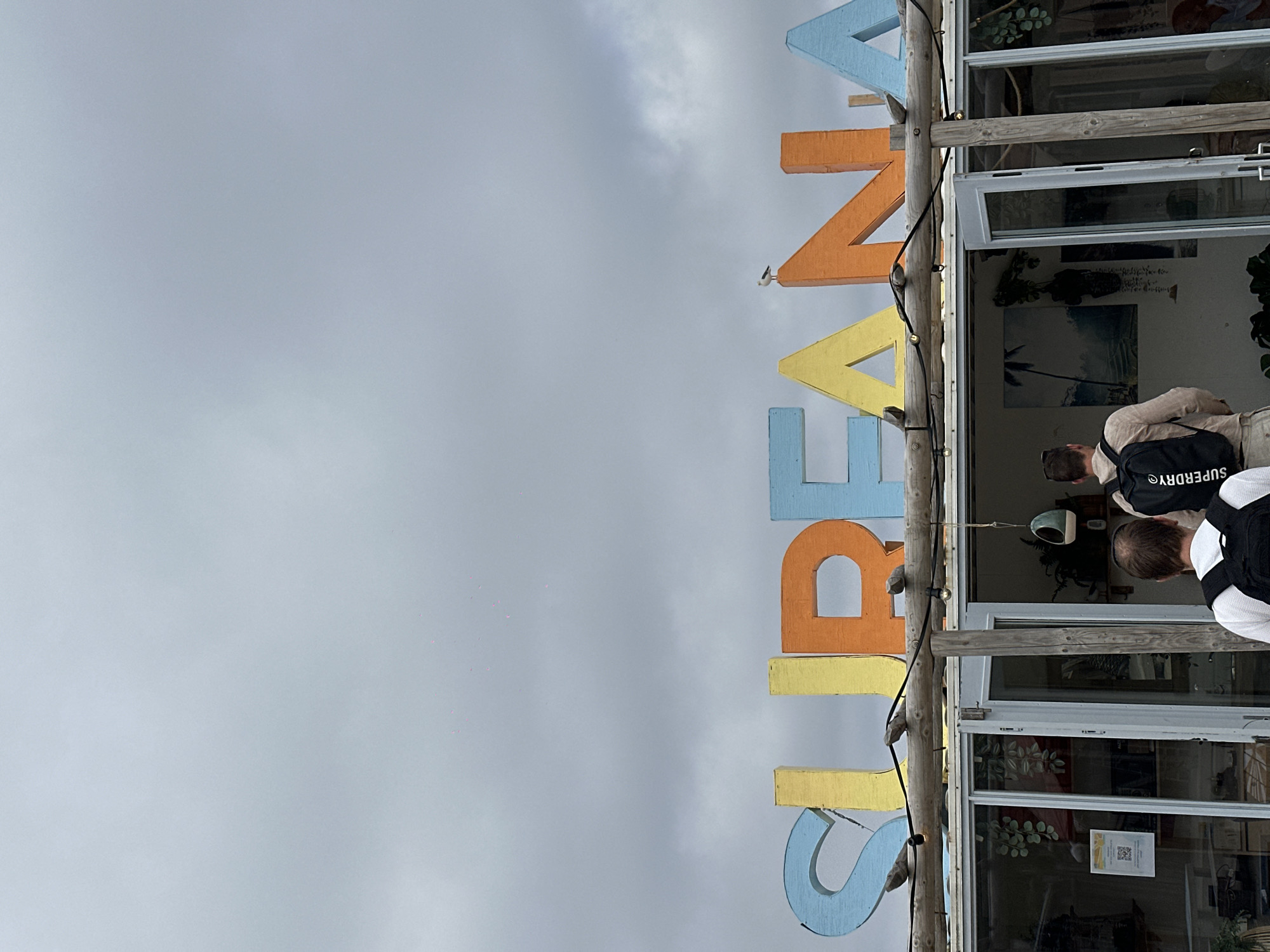 In Overveen, Netherlands, two individuals are captured in a photograph from behind, facing a building adorned with large, colorful wooden letters spelling "SURFANA." The letters, in shades of blue, yellow, and orange, are positioned above the entrance of a store. A seagull is perched atop the letter "N." The building features glass doors and windows, offering glimpses of greenery and artwork within. The sky is overcast with a grey, cloudy atmosphere. One person wears a light brown shirt and a black backpack with "SUPERDRY" visible, while the other is partially visible in the foreground, also wearing a backpack. The overall scene suggests a coastal or surf-themed establishment.
