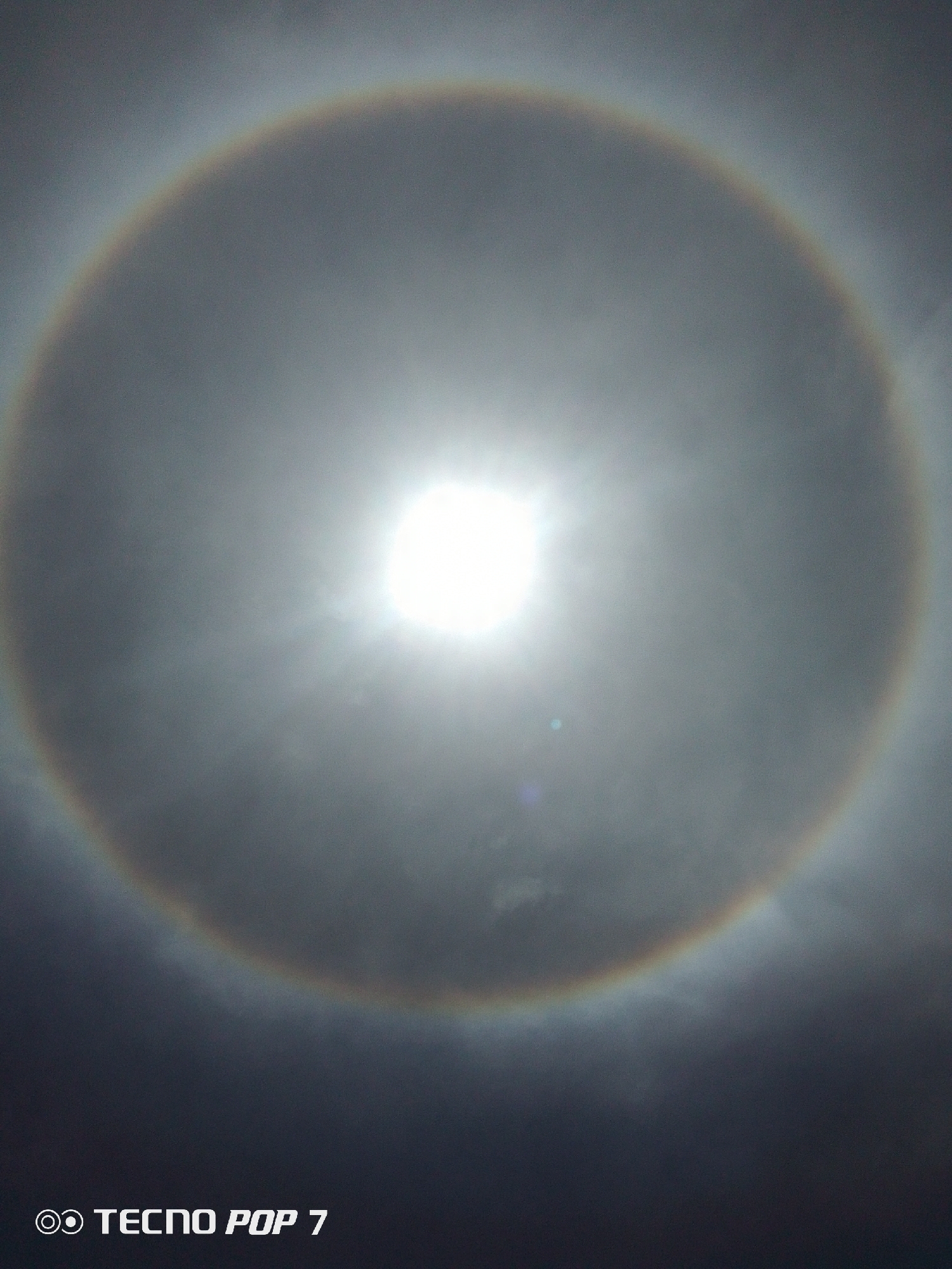 The image depicts a solar halo, a luminous ring around the sun. The halo is visible through a layer of thin clouds, which diffuse the sunlight. The sun appears as a bright white orb at the center of the halo. The circular ring of light is colored in shades of red on the inside and blue on the outside, resembling a rainbow, due to refraction of sunlight by ice crystals in the atmosphere. At the bottom of the image, the words "TECNO POP 7" are visible, indicating the type of phone used to capture the photo.
The photograph was likely taken in Pedro González, Venezuela, as requested. However, no specific information to place this image within that city or its surrounding environment can be determined with this image.