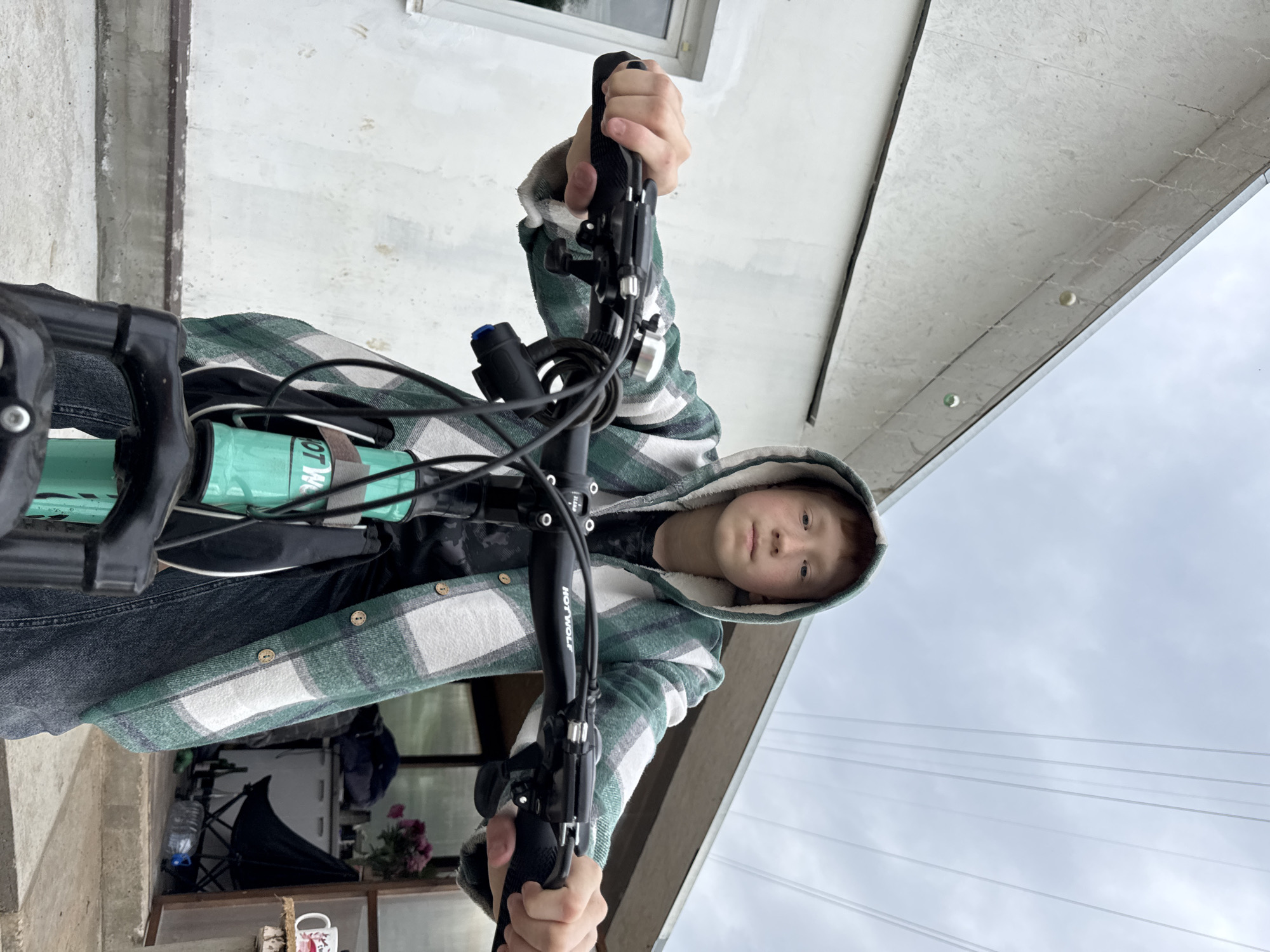 A young person with reddish-blond hair and a serious expression, wearing a green and white plaid hooded jacket over a dark t-shirt and dark jeans, sits on a bicycle. The bicycle has a turquoise frame and black handlebars with the brand name "HOTWOLF" visible. The person's hands are gripping the handlebars. The background shows the side of a white building with a concrete foundation, a window, and overhead power lines against a cloudy sky. To the right, there's an open doorway revealing a cluttered area with chairs, a table, a mug, and bottles. The scene appears to be outdoors, possibly in a rural or suburban setting in Chaikovsky, Russia.