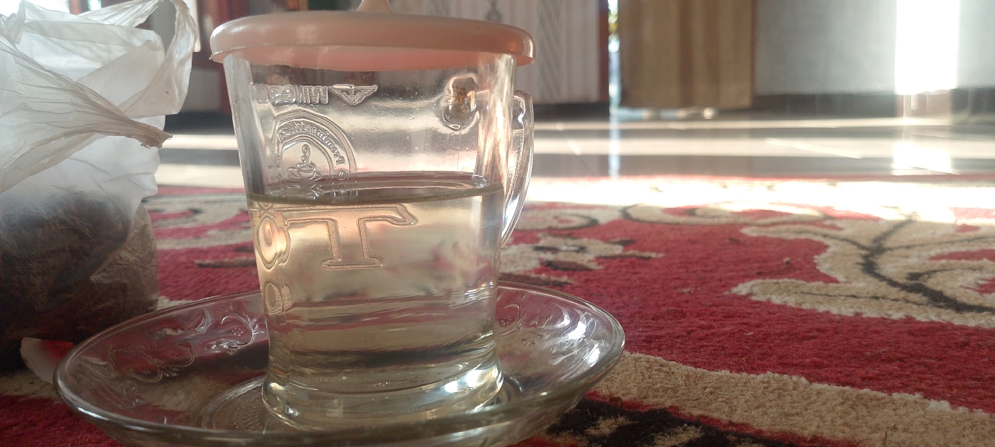 The image shows a close-up, low-angle shot of a clear glass mug filled with a light-colored liquid, likely tea or water, resting on a clear glass saucer. The mug has a pink lid and features embossed designs on its surface, including what appears to be a logo with a steam icon and possibly text. A portion of a white plastic bag containing a brown, granular substance is visible to the left of the mug, with some of the contents spilling out. The saucer and mug are placed on a red carpet with a patterned design in cream and black. In the background, sunlight streams in from the right, illuminating a tiled floor and a blurred view of a doorway or window with drapes. The overall scene suggests a domestic setting, possibly indoors, with natural light being a prominent feature. The image does not provide any information about the specific location in Oro-oro Ombo, Indonesia.