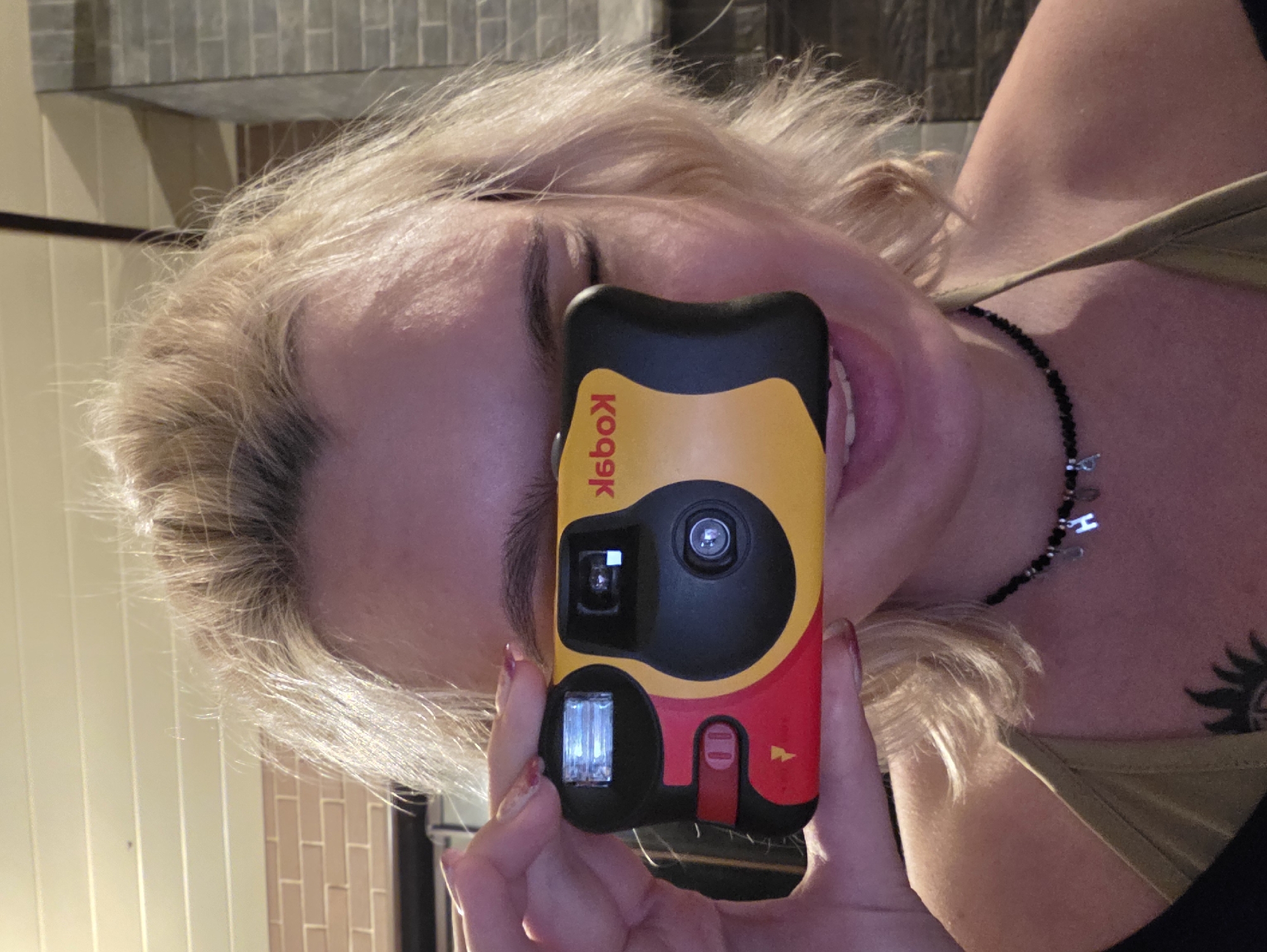 A young woman with blonde, short hair is holding a red and yellow disposable camera up to her face, covering her eyes and nose. She is wearing a tan tank top and a black beaded necklace with a silver charm. The background shows a brick wall and a white ceiling with horizontal lines. The scene is indoors, likely at night given the lighting conditions. There is no indication of location in the city of Нагорный, Russia.