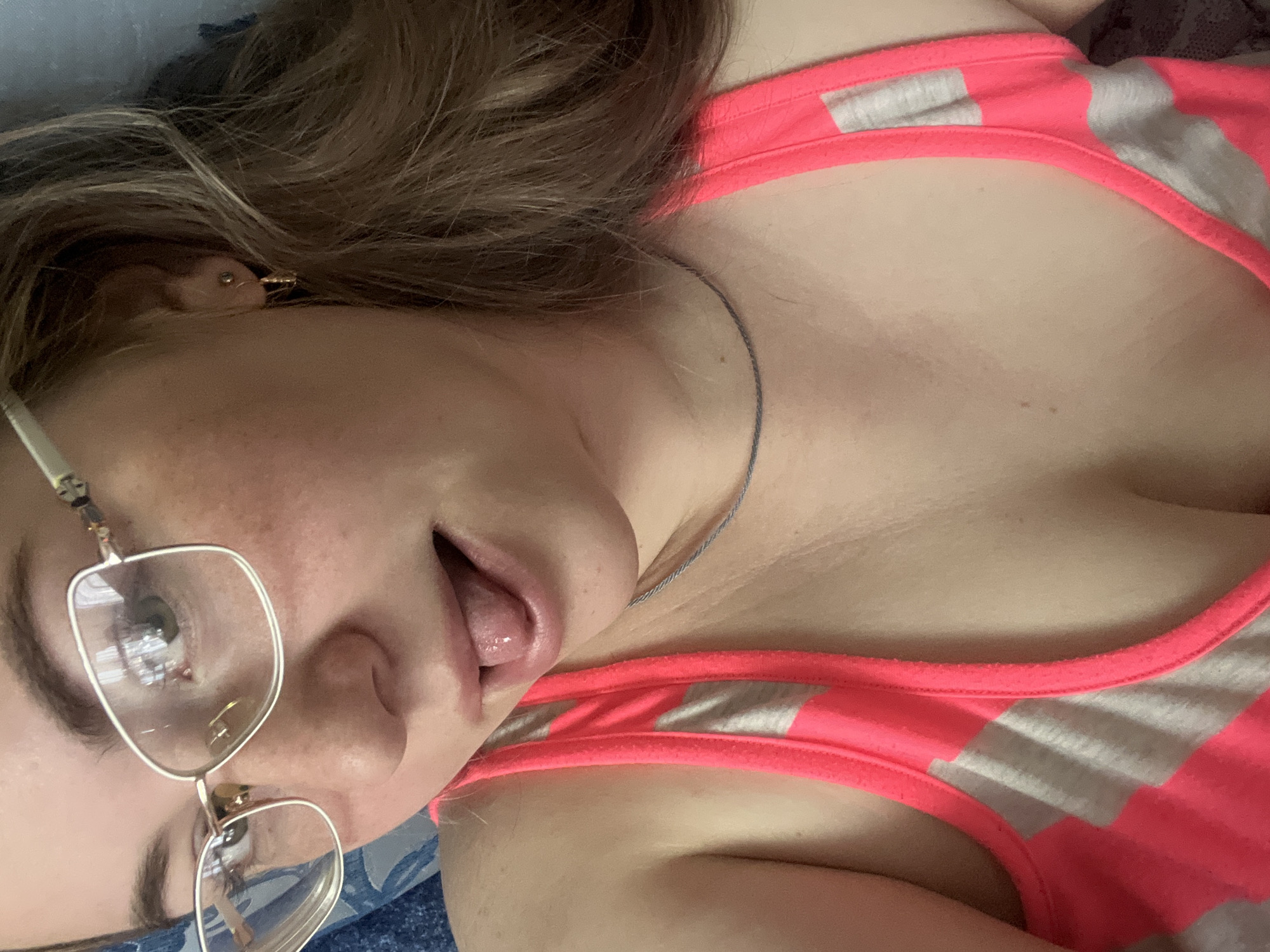 A young woman with light skin, wearing gold-rimmed glasses and a pink and grey striped tank top, is lying on a surface with a blue floral pattern. Her head is tilted, and she has her tongue out. A thin silver necklace is visible around her neck, and she has an earring in her left ear. Her hair is brown. The location is not discernible from the image. The scene does not appear to be located in Chaikovsky, Russia, due to the lack of discernible landmarks.