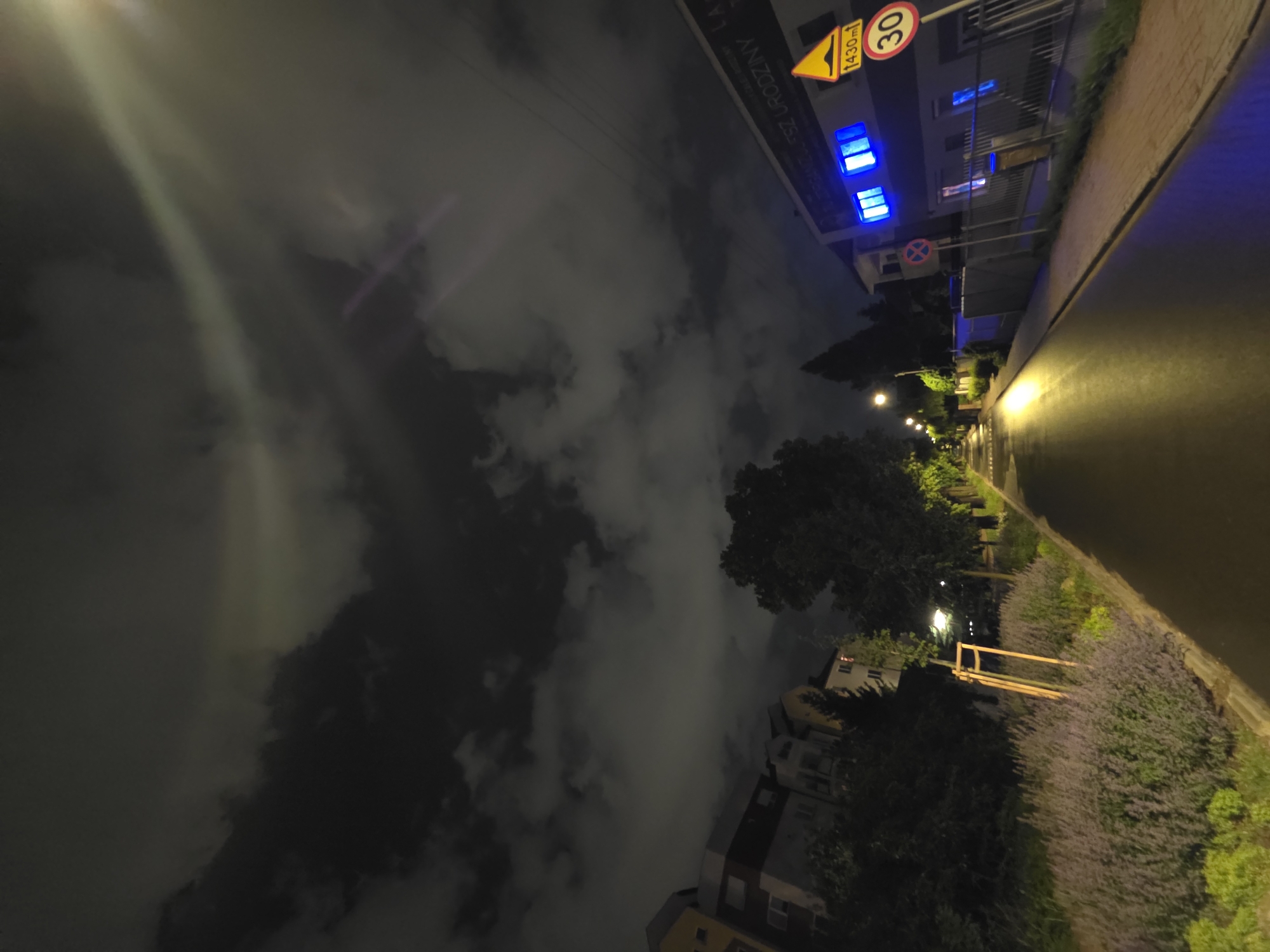 Night shot in Warsaw, Poland. The view is along a street with a sidewalk and buildings on either side. Streetlights illuminate the road. A "30" speed limit sign and a triangular hazard sign are visible on a building. Clouds dominate the sky.
