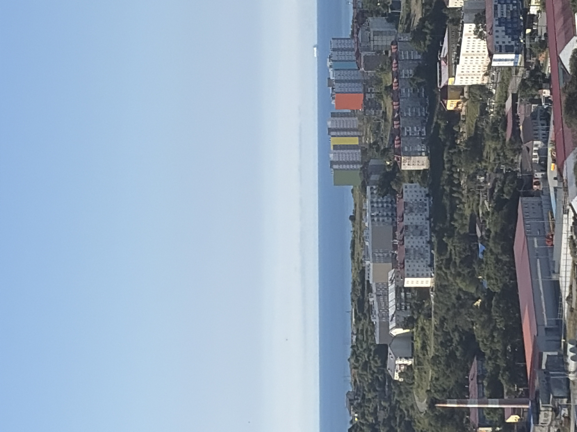 The image is a high-angle shot of the city of Korsakov, Russia, showing a densely populated area with buildings situated on a hillside overlooking the sea. In the foreground, a large industrial building with a red roof is visible, along with a tall, striped smokestack. Further up the hill, residential buildings of various sizes and architectural styles are clustered together, some with colorful facades. The background features a vast expanse of blue sea under a clear sky, with a distant ship visible on the horizon. The overall impression is of a working-class city with a mix of industrial and residential areas, set against a scenic coastal backdrop.
