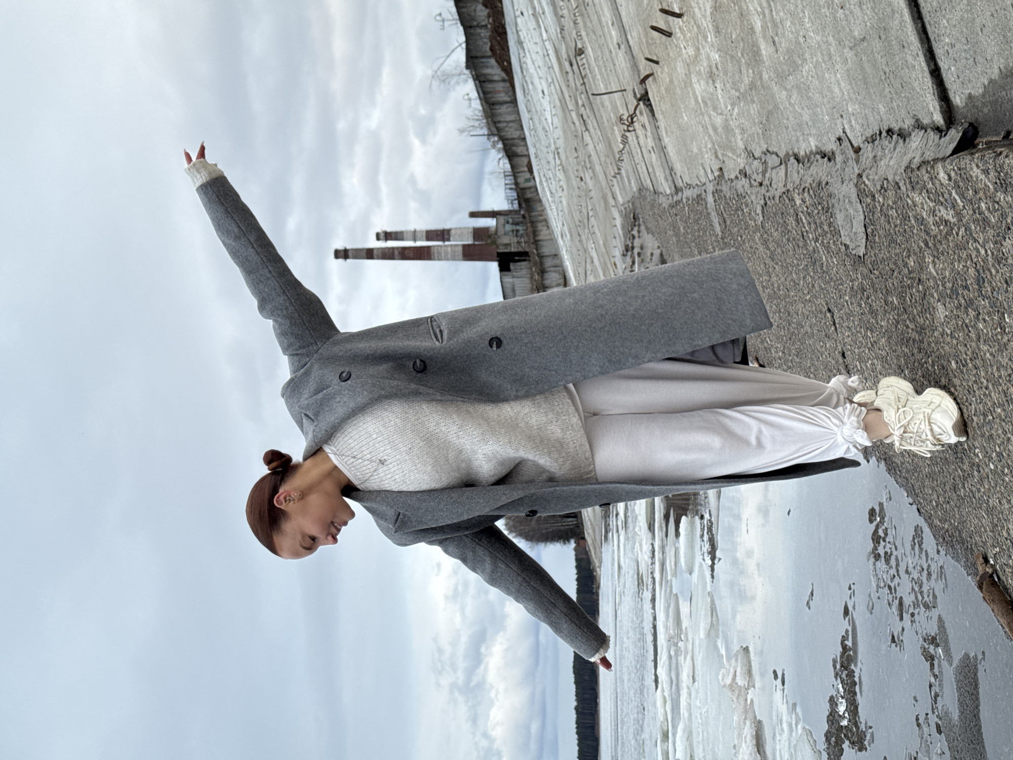 A young individual, with light brown hair neatly styled in a bun, strikes an expressive pose outdoors, captured in a full-body shot. They are dressed in a long, dark grey overcoat, a light-colored sweater, matching light-colored pants, and chunky white sneakers, indicating cold weather. Their left arm is extended high and outward towards the sky, with fingers slightly apart, while their right arm is extended downwards and behind them, creating a dynamic, balanced posture. The person's head is tilted downwards, and they appear to be looking towards their left.
The background features a vast, overcast grey sky. Below the sky, two tall, striped industrial smokestacks are visible in the distance, suggesting a nearby factory or power plant. In the midground, a worn concrete or stone retaining wall runs parallel to the picture plane. To the right, a rough, textured concrete surface forms the immediate ground. The foreground and midground on the left reveal a body of water largely covered in melting ice chunks and slush, characteristic of a cold, possibly late winter or early spring environment. The scene, with its blend of industrial elements and a partially frozen landscape, aligns with locations like Chaikovsky, Russia. No visible text is present in the image.