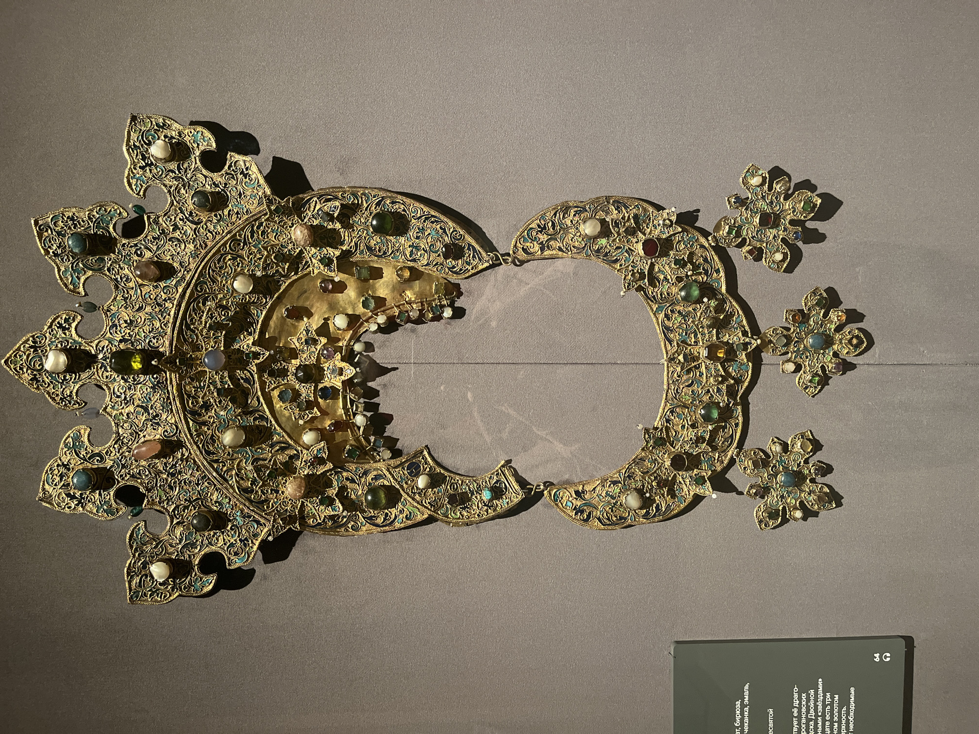 The image shows a decorative gold artifact displayed against a plain gray backdrop. The artifact is intricately designed with a semi-circular structure, adorned with various colored stones and detailed filigree work. Its overall shape resembles a stylized crown or headpiece. The artifact appears to be in two separate sections, with a gap between them. Several smaller, flower-like ornaments are attached or positioned nearby, complementing the design. The artifact is possibly on display in a museum, given the plain backdrop and a sign with text in Cyrillic in the lower right corner. The setting is Unknown in Unknown, Unknown.