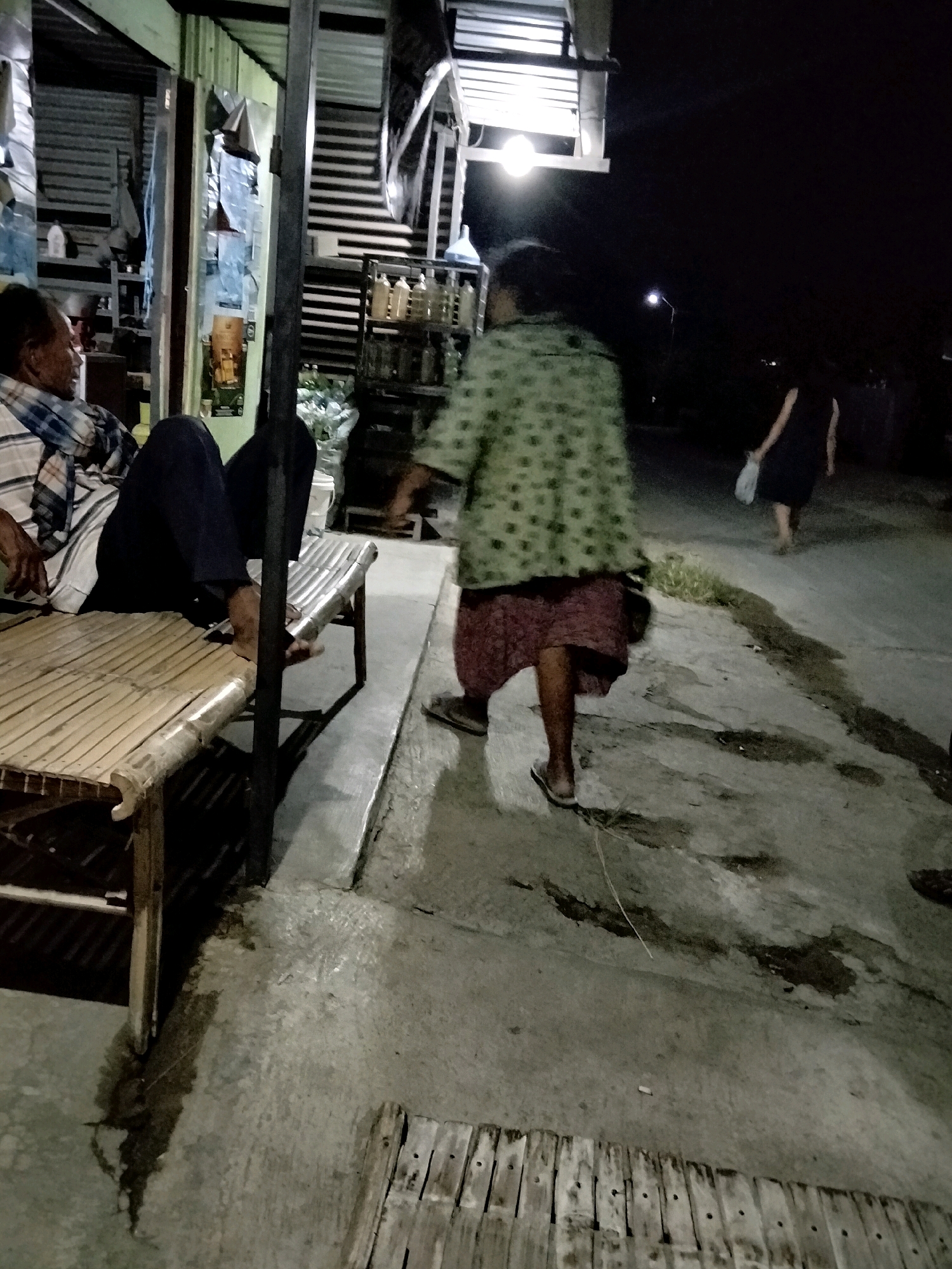 In Mrican, Indonesia, a man is seated on a bamboo bench, facing left, with his legs extended. He is wearing a plaid shirt and dark pants. To his right, a portion of another bamboo bench is visible. Further in the background, a woman wearing a green patterned jacket and a reddish-brown skirt walks away from the camera. Another woman, dressed in a dark garment, is walking in the distance, holding a white bag. The scene takes place at night, with artificial lighting casting shadows on the concrete ground. Structures with corrugated metal siding and shelves with bottles are visible in the background.