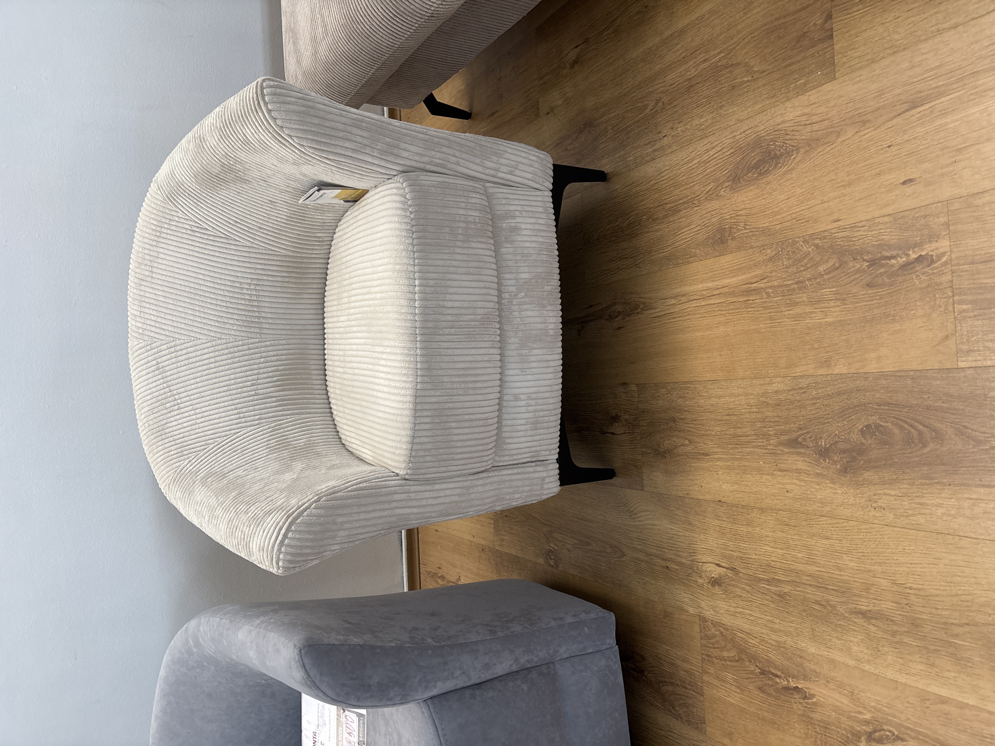 The image shows two pieces of furniture in an indoor setting, likely a living room or showroom. The primary subject is a cream-colored armchair with a corduroy texture. It has a rounded, enveloping design and sits on black angular legs. A small yellow tag is visible tucked into the armrest. Next to it, partially in the foreground, is a grey upholstered ottoman or stool, also with a soft texture and curved lines. The flooring is made of light brown wood planks with a visible grain pattern. The background features a light blue wall. The lighting appears to be artificial, suggesting an indoor environment, and there are no indications of the time of day or weather. The scene is static, with no people or discernible activity taking place. The overall impression is one of modern furniture displayed in a clean, minimalist space. There is no discernible text in the image.