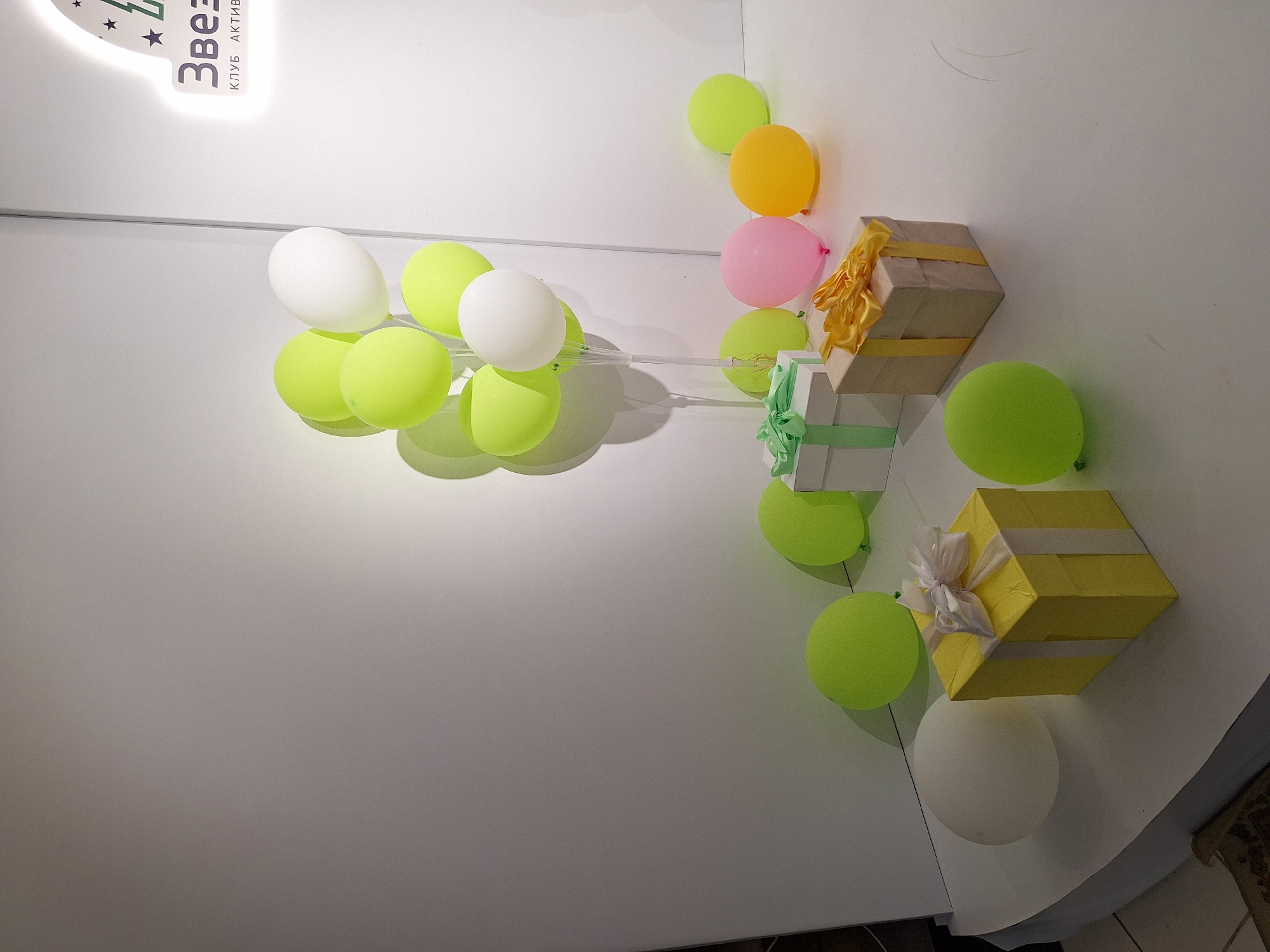 The image depicts a celebratory scene, possibly a party or event, set against a plain white background. A cluster of lime green and white balloons are suspended from clear plastic stands in the center of the composition. Scattered around the balloons are several gift boxes, adorned with ribbons. One box is yellow with white ribbon, another is white with green ribbon, and a third appears to be kraft paper with yellow ribbon. There are also individual balloons in lime green, pink, and yellow on the floor. A logo with the word "Звезд" (Stars) in Cyrillic script and small star graphics is visible in the upper right corner, suggesting the event might be taking place at a venue named "Stars Club." The overall impression is one of a festive and organized event. The location is the city of Атамановка, Russia.