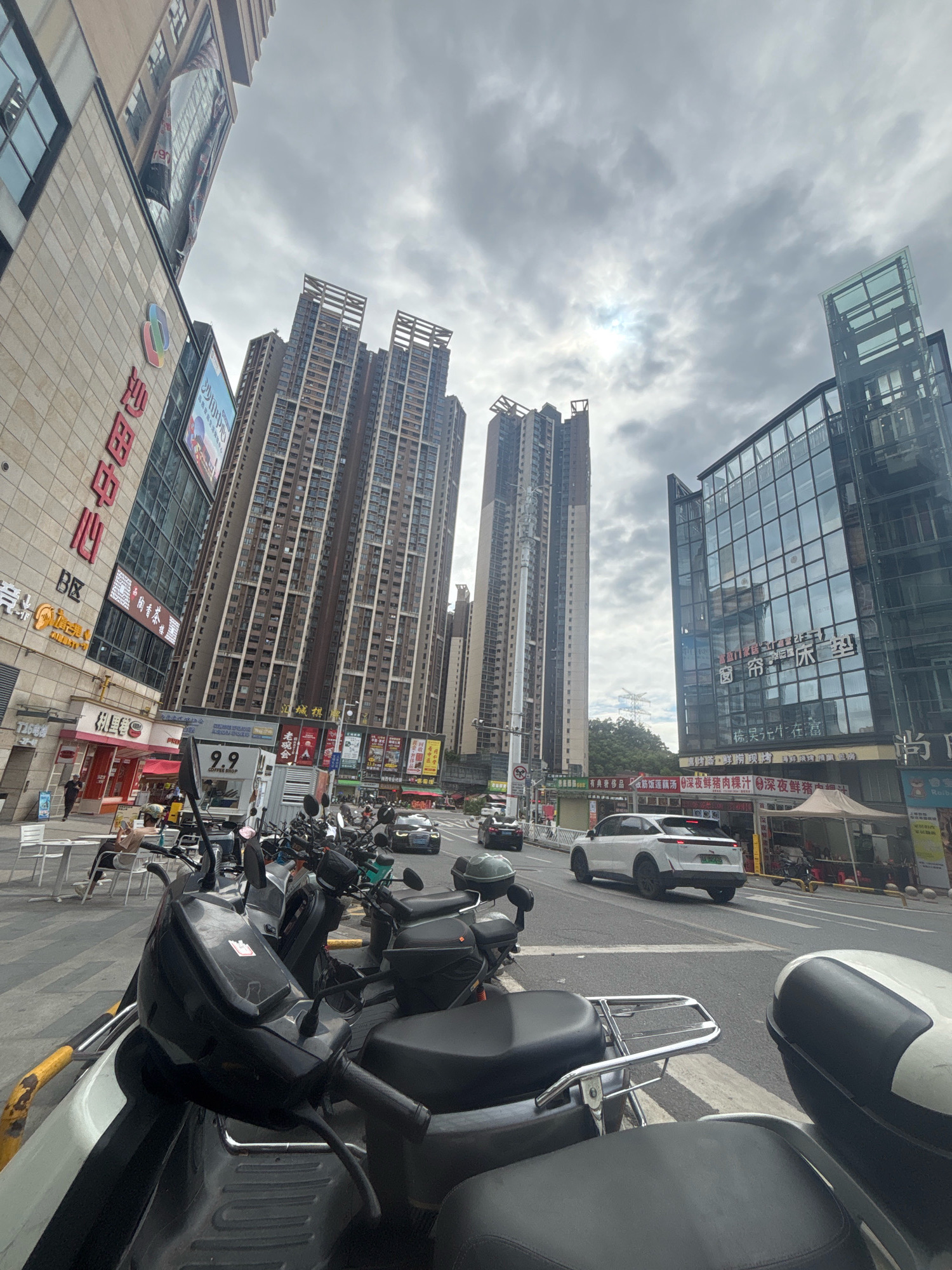 The image captures a street scene in Nanhai District, China, characterized by modern urban architecture and a cloudy sky. In the foreground, several parked electric scooters and motorcycles are visible, suggesting a common mode of transportation. The street itself is paved, with yellow and black striped barriers indicating pedestrian crossings or traffic dividers.
Dominating the background are towering apartment buildings and commercial structures. On the left, a beige building with red Chinese characters and a colorful logo, likely a commercial establishment, is prominent. Adjacent to it, a much taller, slender high-rise apartment building with numerous windows is situated. Further to the right, another imposing apartment building, wider and with a distinctive top section, stands tall. On the far right, a modern building with a glass facade and blue architectural accents reflects the cloudy sky.
Several cars are present on the road, with one white SUV prominently featured in the middle ground. Other vehicles are partially visible, indicating moderate traffic. The street level also shows storefronts, awnings, and signage, hinting at a commercial district. Small white tables and chairs are set up on the sidewalk on the left. The overall impression is of a bustling urban environment under an overcast sky, typical of a developed city in China.