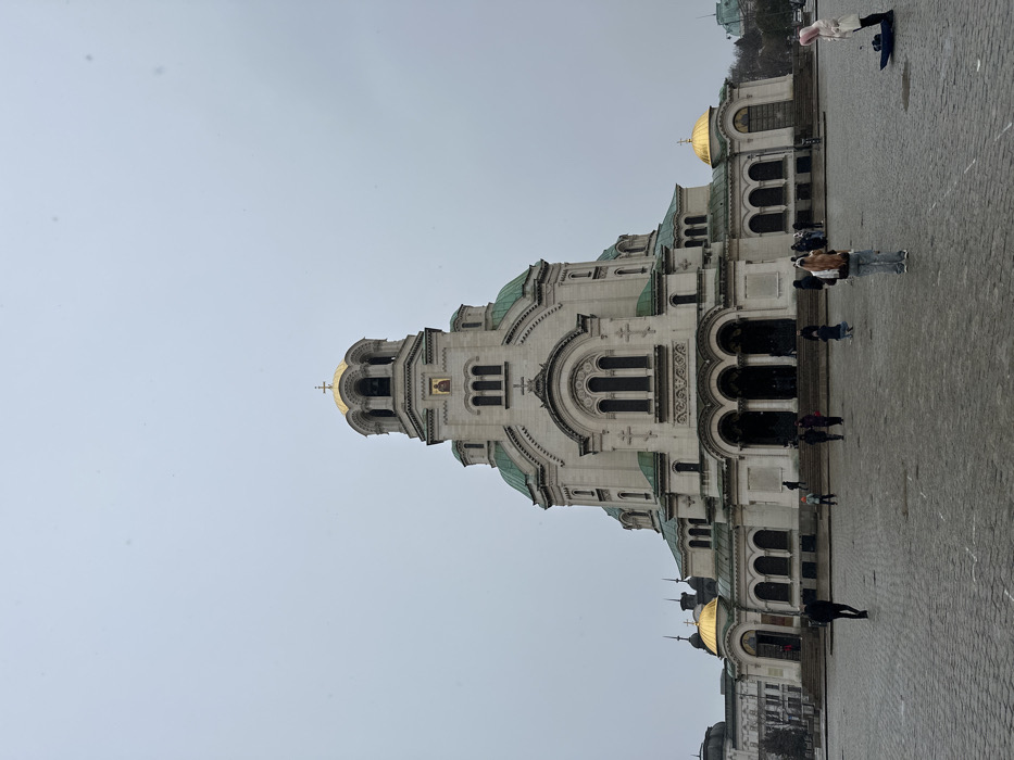The image depicts the Alexander Nevsky Cathedral in Sofia, Bulgaria, under an overcast sky. The prominent subject is the large Orthodox cathedral, constructed of light-colored stone with multiple green domes and spires, and a central golden dome crowned by a cross. A smaller golden dome is visible on the left side of the cathedral's facade. The building features arched windows, ornate detailing, and a central entrance with multiple arches and steps leading up to it. A religious icon is visible on the central bell tower below its golden dome.
The foreground consists of a wet, light-colored cobblestone plaza. Approximately 15-20 individuals are present in the plaza and on the cathedral steps. In the mid-ground, a person with long, light-colored hair, wearing a light-colored top and blue jeans, stands facing the cathedral. To their right, another individual with pink hair, dressed in a white coat, stands near a dark object on the ground. Other people are dispersed across the plaza, walking or standing; some are ascending the cathedral steps. A multistory building with a darker dome is partially visible on the far left behind the cathedral. The overall atmosphere is dim due to the gray sky.